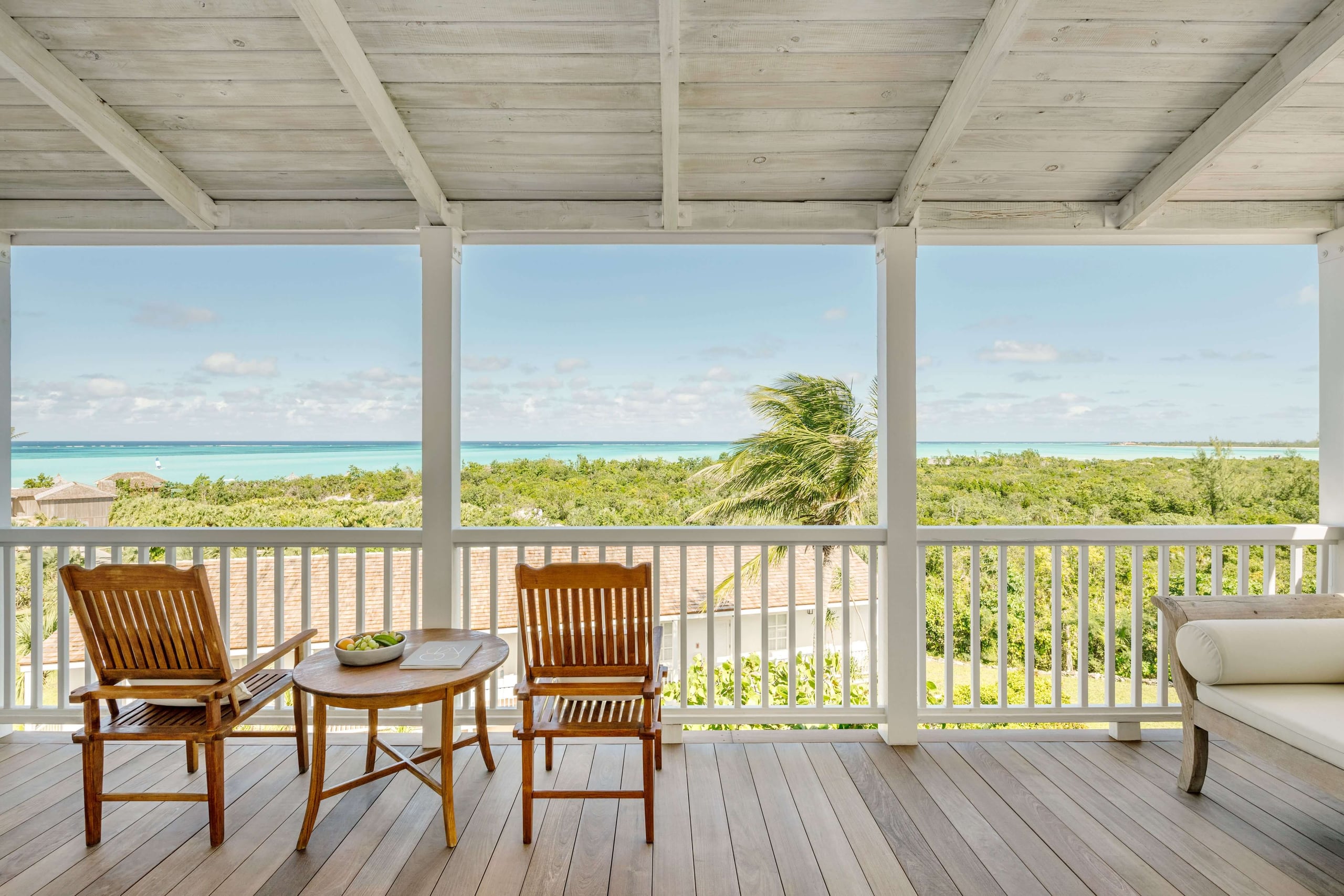 COMO Parrot Cay, North America — Oceanfront terrace with teak lounge chairs, wooden beamed ceiling, turquoise waters and lush vegetation beyond white railing.