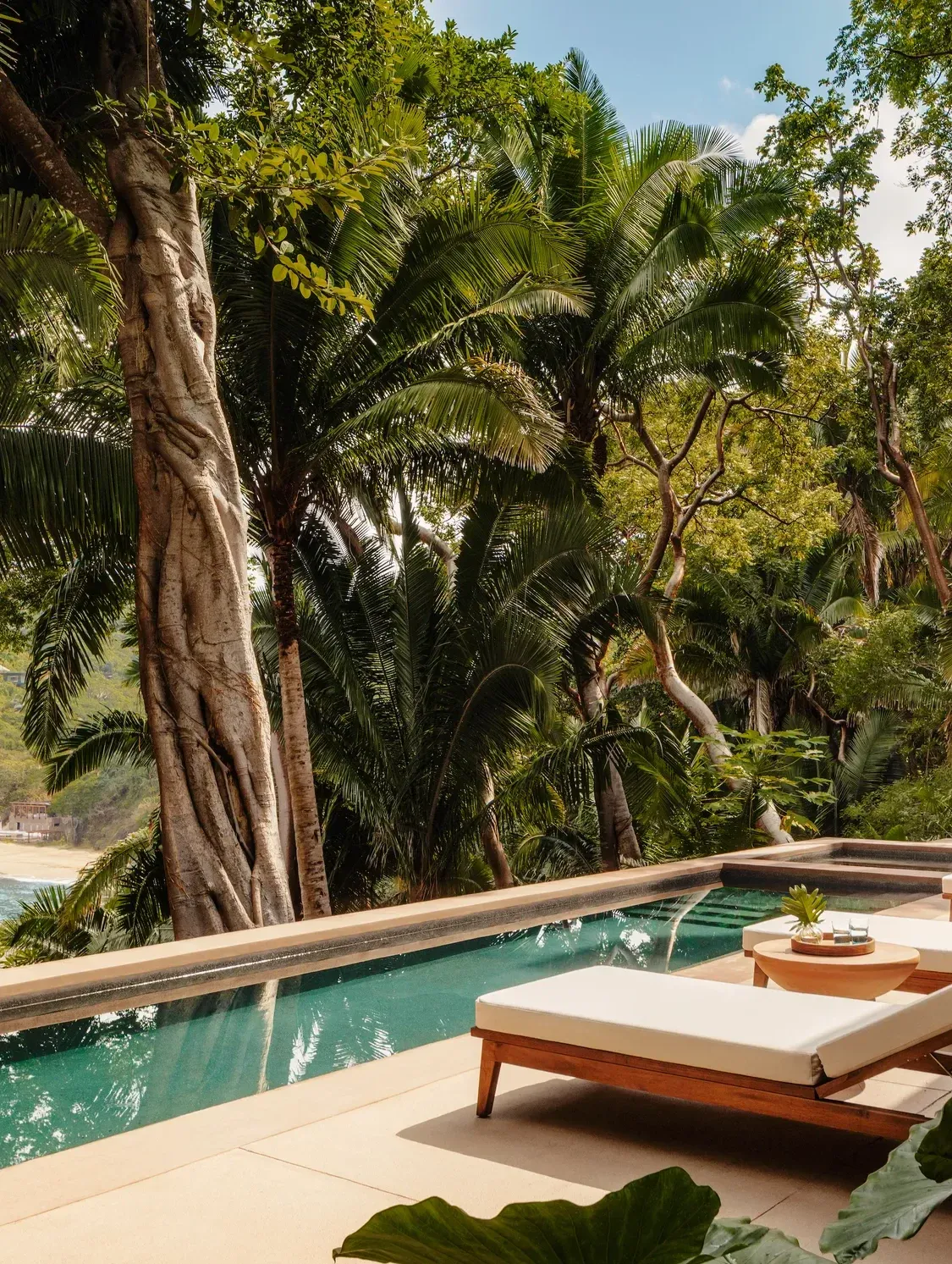 One&Only Mandarina, Mexico — Infinity pool with turquoise water overlooks tropical beach, flanked by towering palms and hardwood lounger under dappled light.
