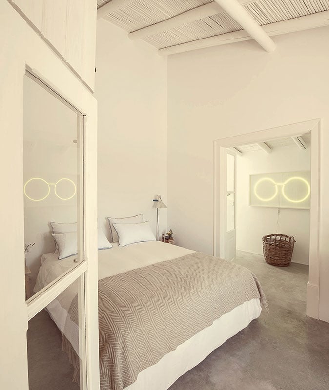 Pensão Agrícola, Europe — Minimalist bedroom with neon circle art, polished concrete floors, neutral linens, and open doorway to ensuite bathroom with natural light.