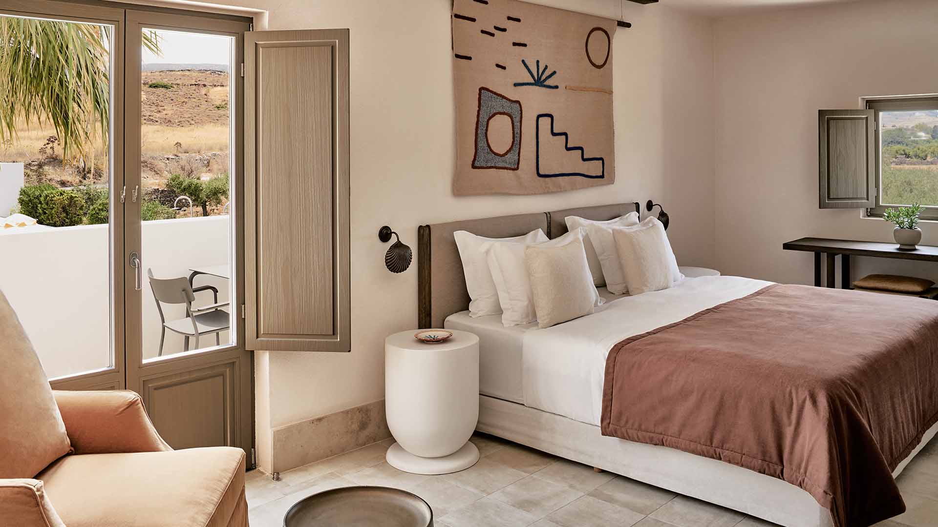 Parīlio Hotel, Europe — Minimalist bedroom with terracotta bedding, cream pillows, curved white nightstand, abstract wall tapestry, and desert landscape visible through glass doors.