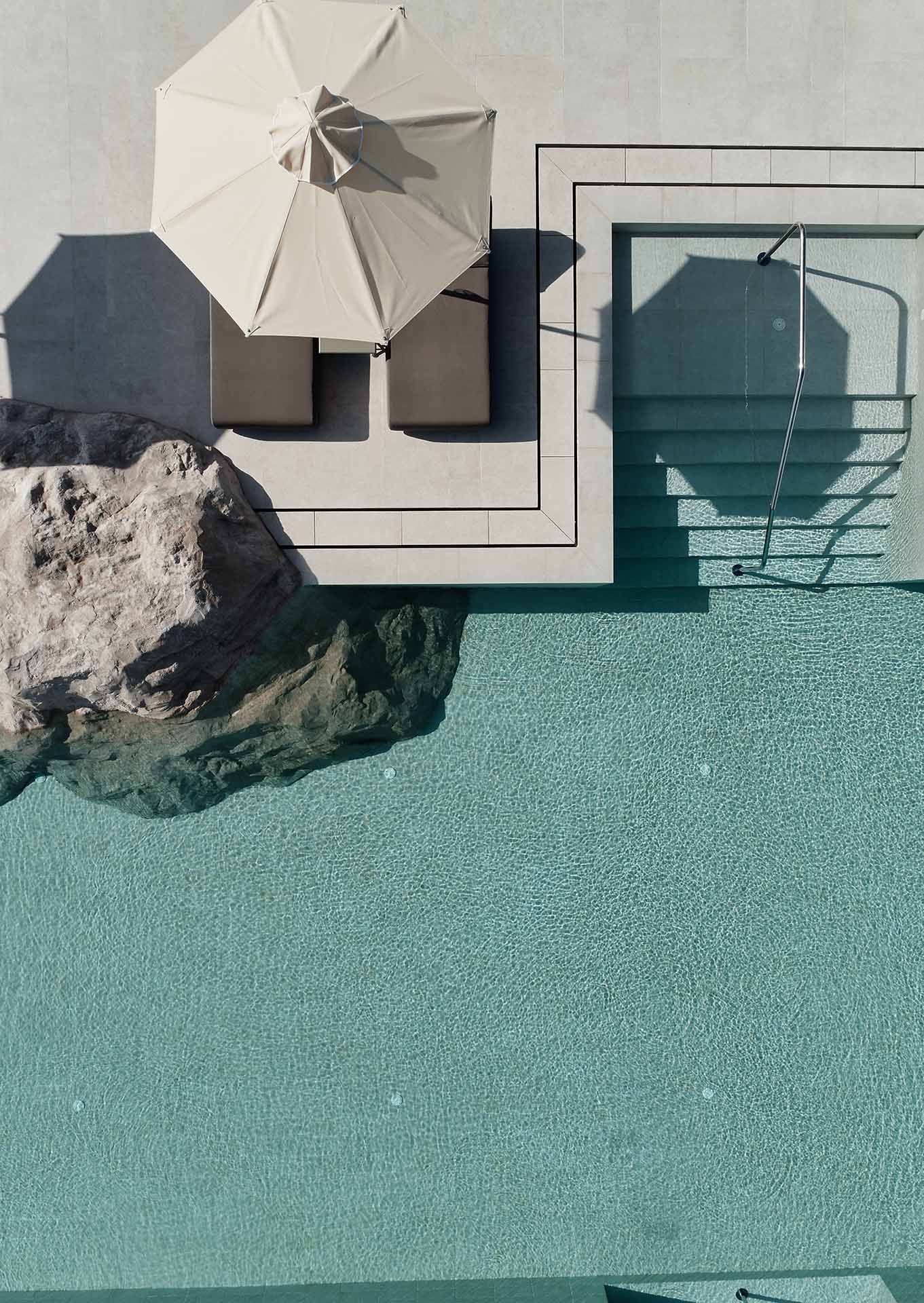 Parīlio Hotel, Europe — Minimalist poolside terrace with cream umbrella, turquoise water, natural stone, and modernist architecture casting geometric shadows.