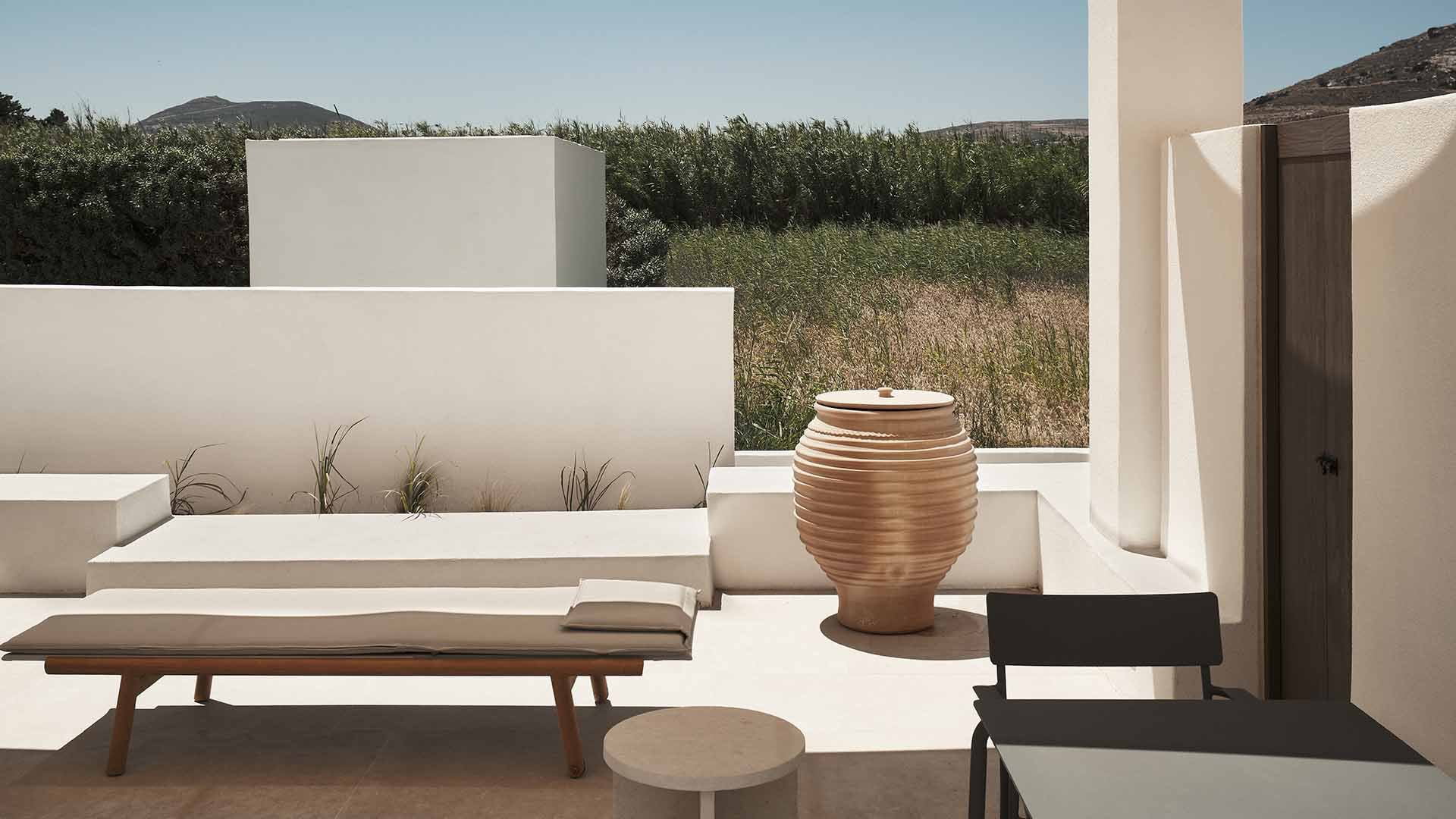 Parīlio Hotel, Europe — Minimalist courtyard with white concrete walls, terracotta vessel, wooden bench, and olive groves beyond, bathed in Mediterranean sunlight.