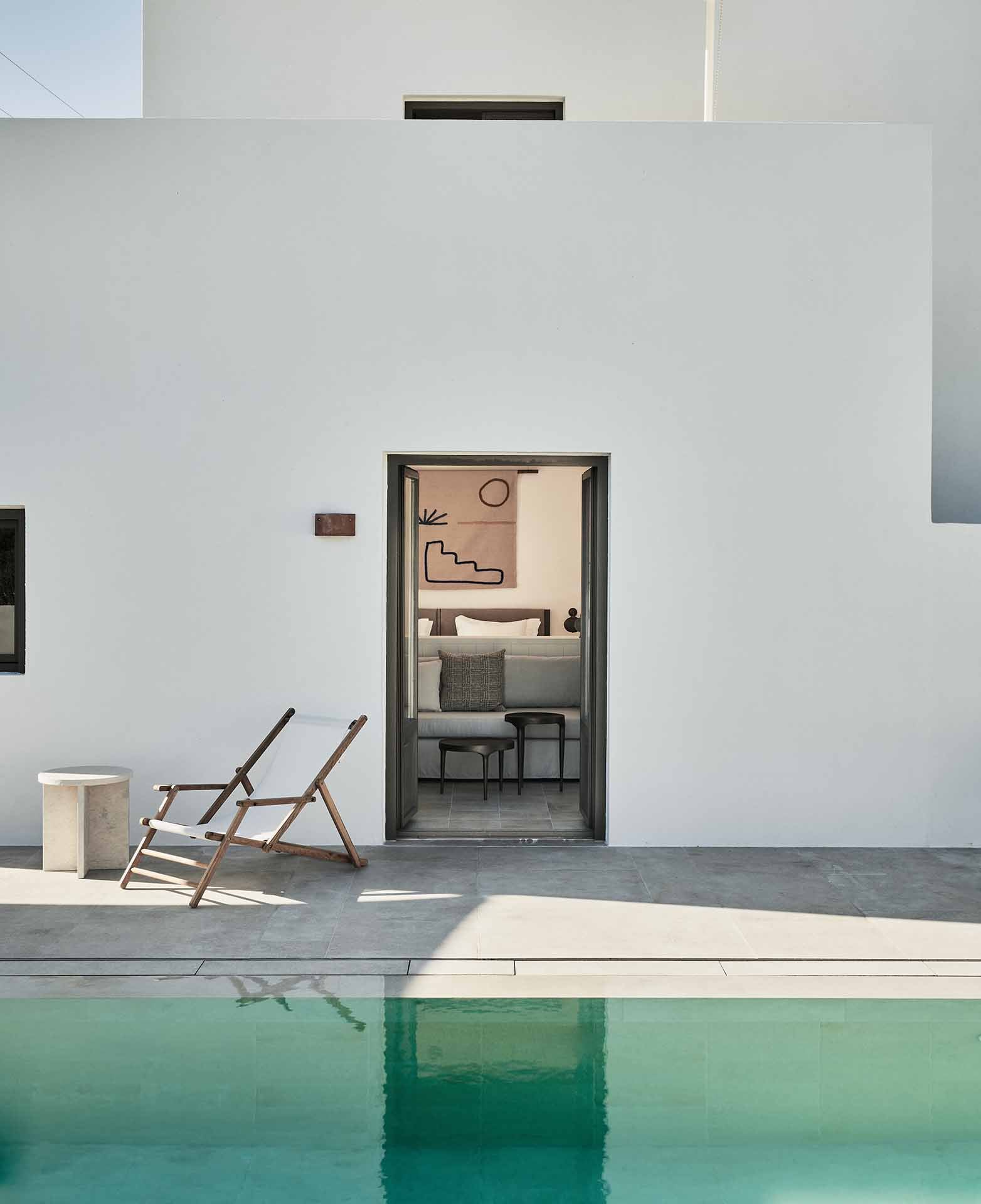 Parīlio Hotel, Europe — Minimalist poolside bedroom with whitewashed walls, wooden lounge chair, and turquoise water reflecting modernist architecture.