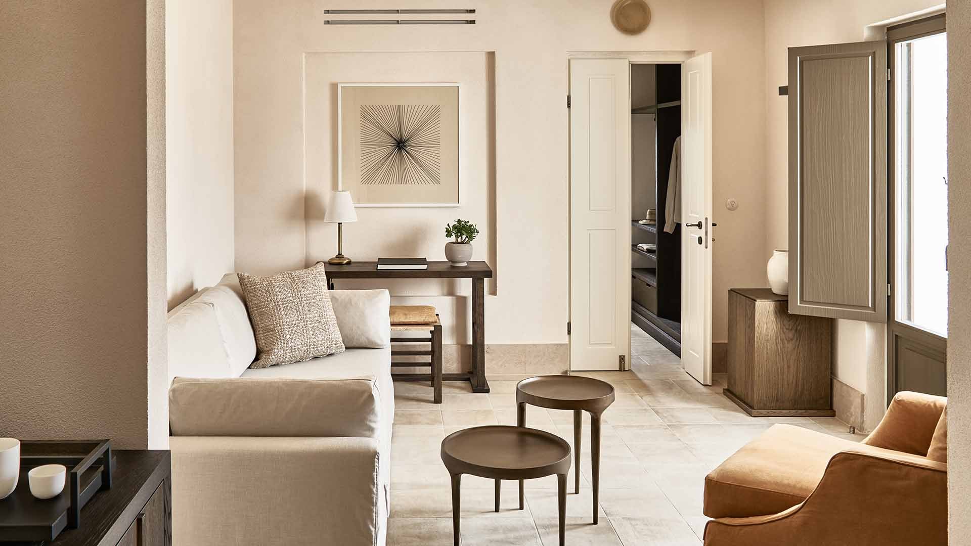 Parīlio Hotel, Europe — Minimalist suite with cream linens, walnut side tables, bronze accent chairs, and soft natural light filtering through glass doors.