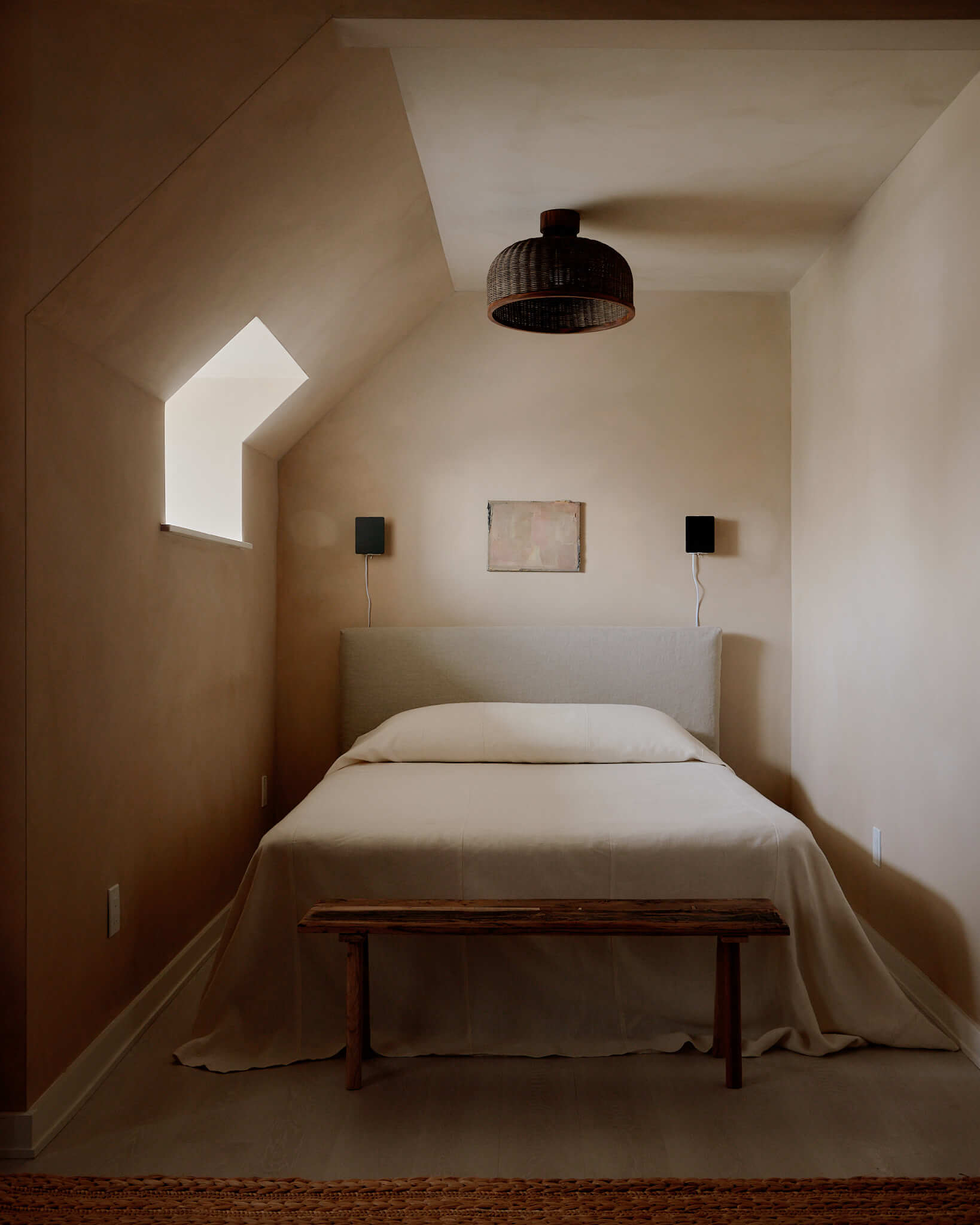 The James Bradley, North America — Minimalist bedroom with sloped ceiling, skylight flooding natural light, rattan pendant fixture, cream bedding, and wicker speakers on neutral walls.