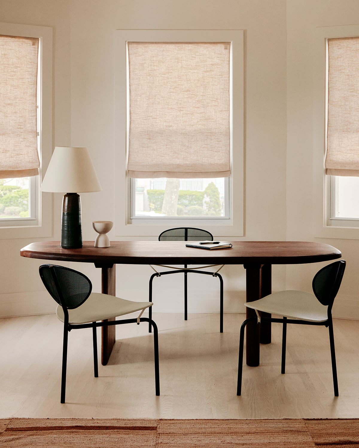 The James Bradley, North America — Minimalist dining room with oval walnut table, black metal chairs, linen roman shades, and warm natural light from multiple windows.