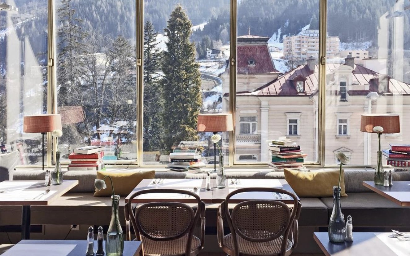 Regina Bar & Restaurant, Europe — Long wooden desks with bentwood chairs overlooking snow-capped Alpine mountains, forested valleys, and Belle Époque architecture through floor-to-ceiling windows.