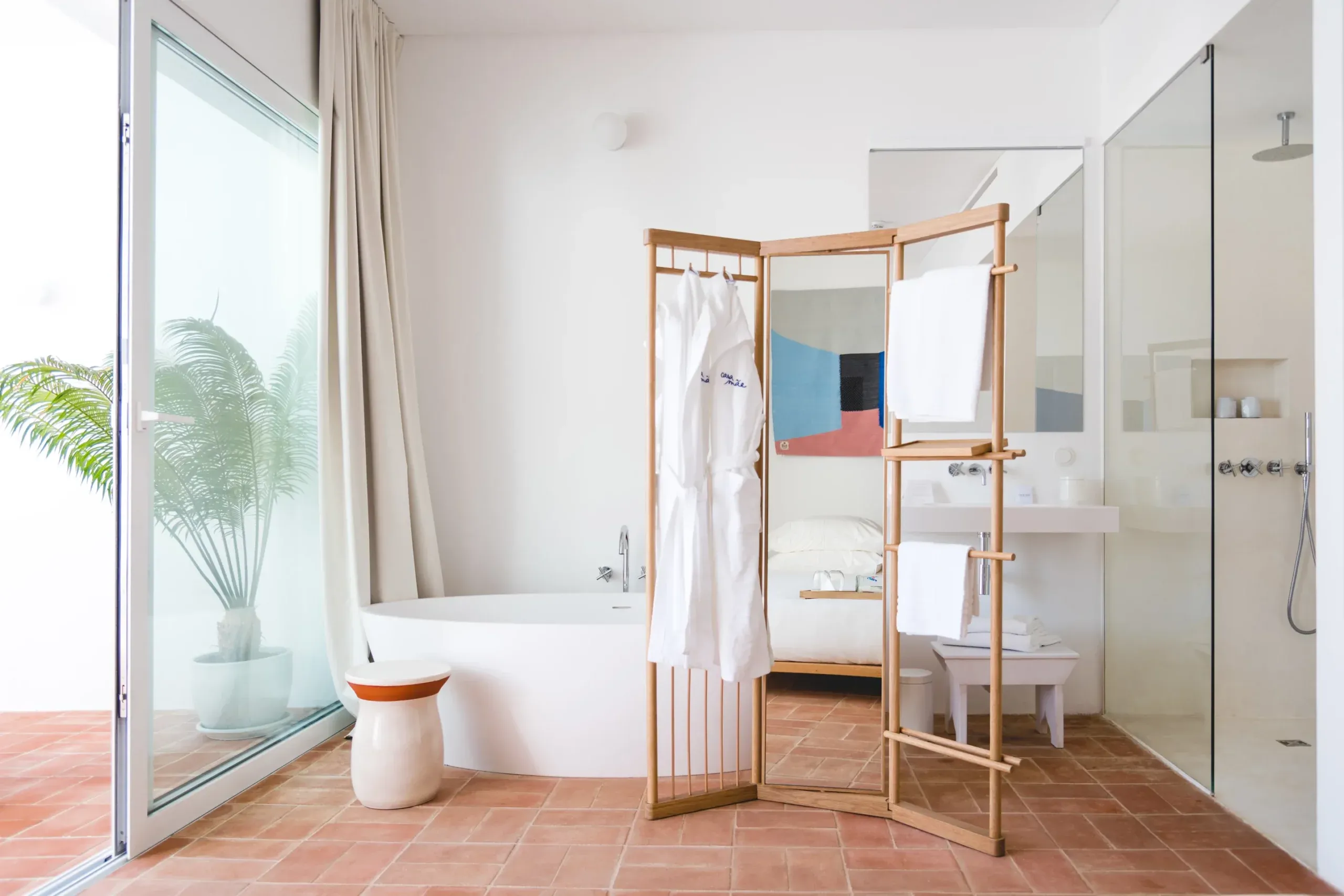 Casa Mãe, Europe — Bathroom with curved white soaking tub, wooden clothing rack, terracotta tile floor, glass shower enclosure, and bedroom visible through open doorway.