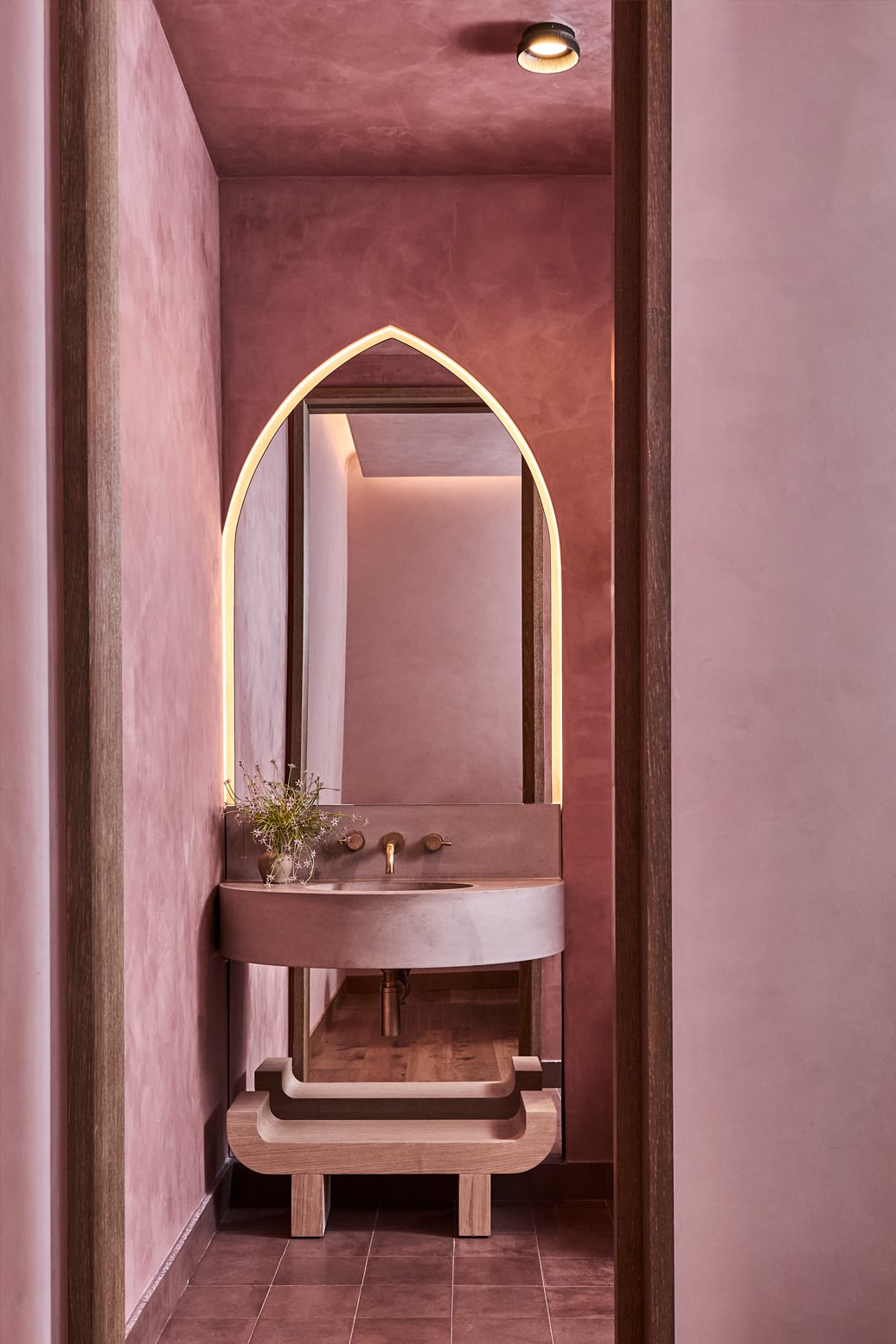 Surya Spa, North America — Arched mirror with warm ambient lighting above curved ceramic sink in mauve plaster bathroom with wood accents.