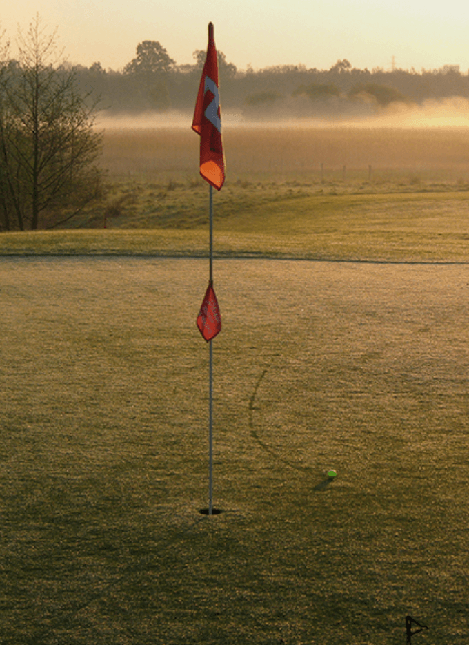 Golf de Rochefort, Europe — Red flag marking golf hole on misty morning fairway with frost-covered grass and tree line.