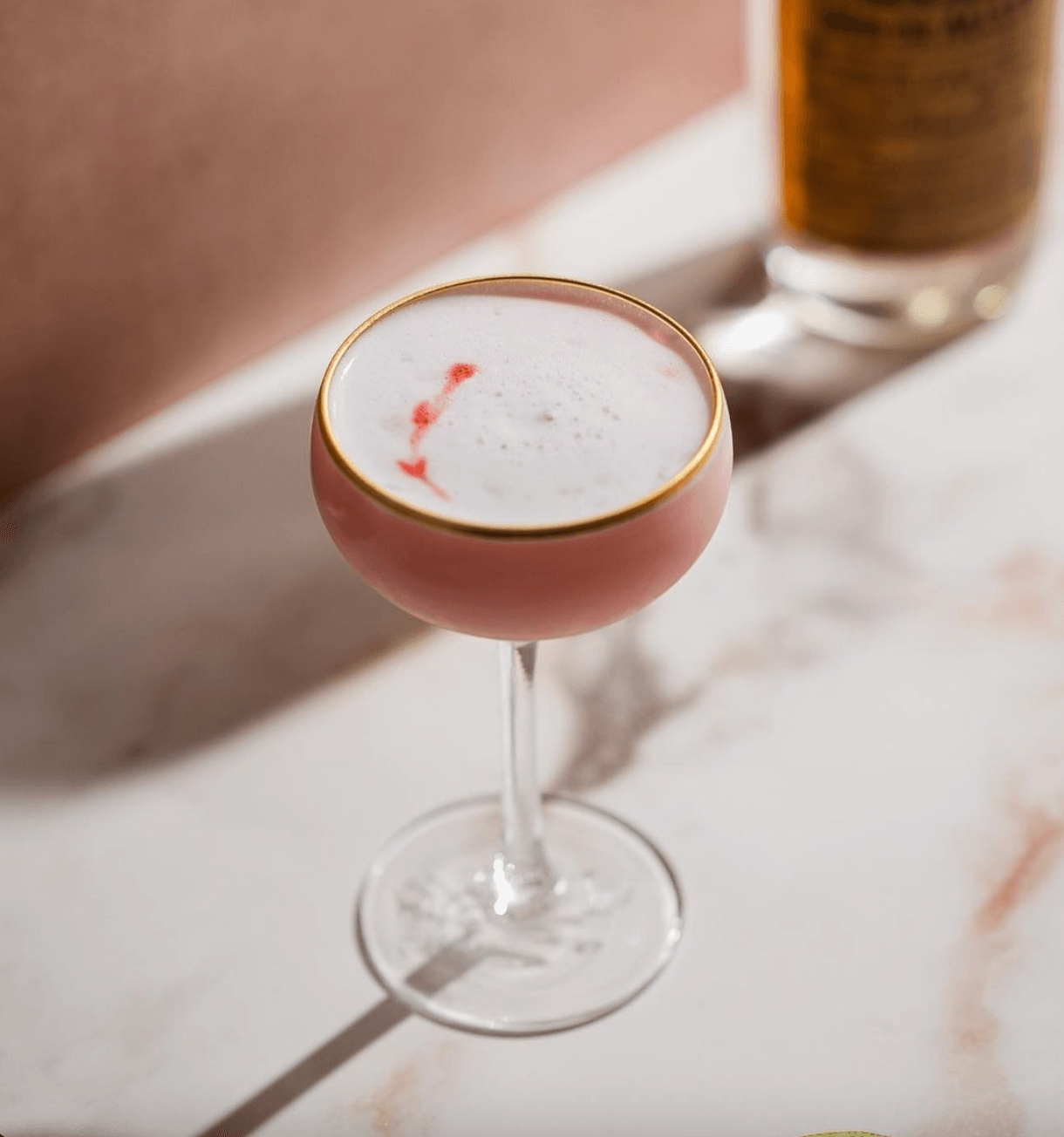 Death & Co, North America — Pink cocktail with gold rim and red bitters detail in coupe glass, minimalist bar setting with marble surface.