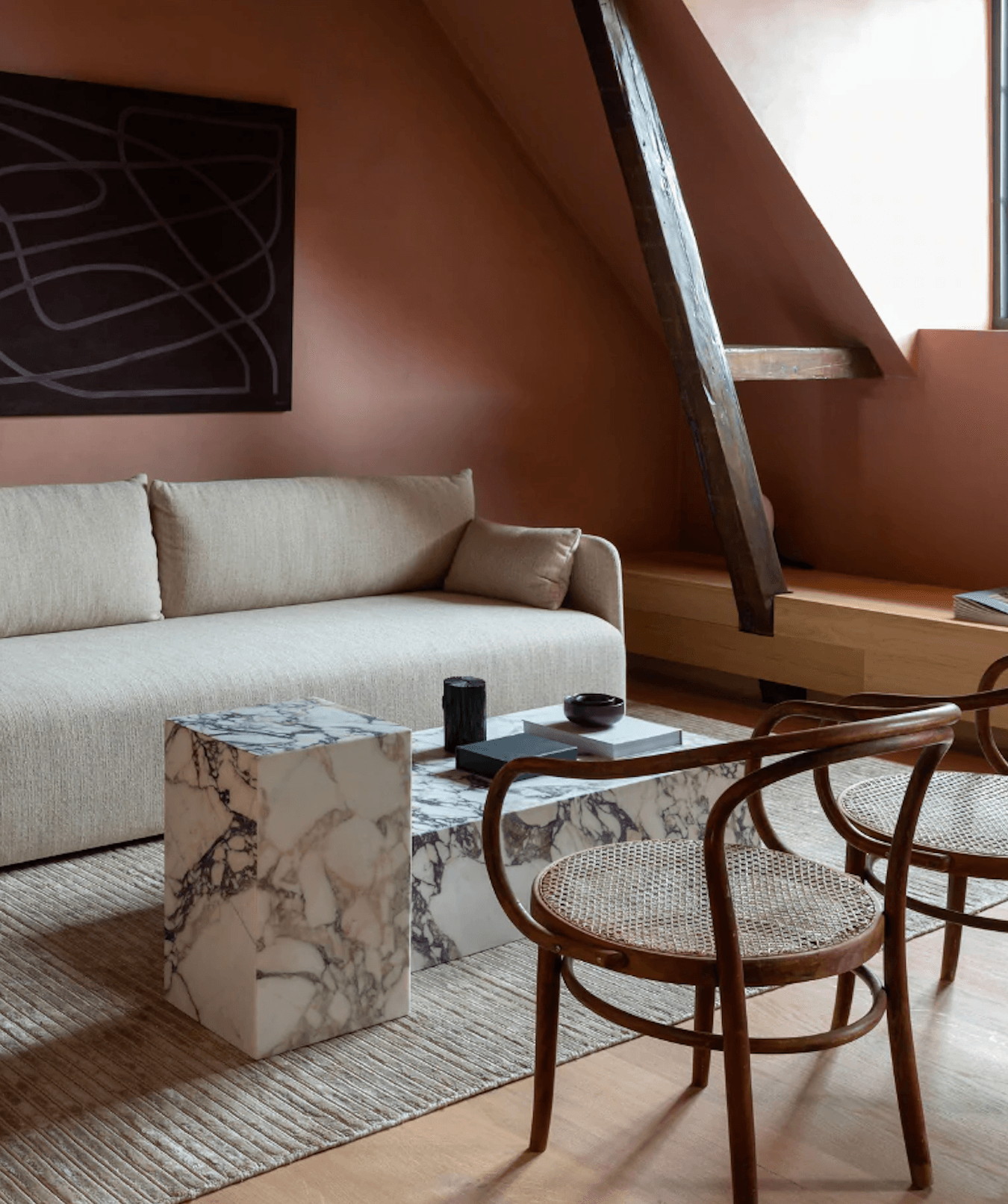 The Audo, Europe — Minimalist attic suite with terracotta walls, cream linen sofa, marble side table, bentwood chairs, and sculptural timber staircase in soft natural light.