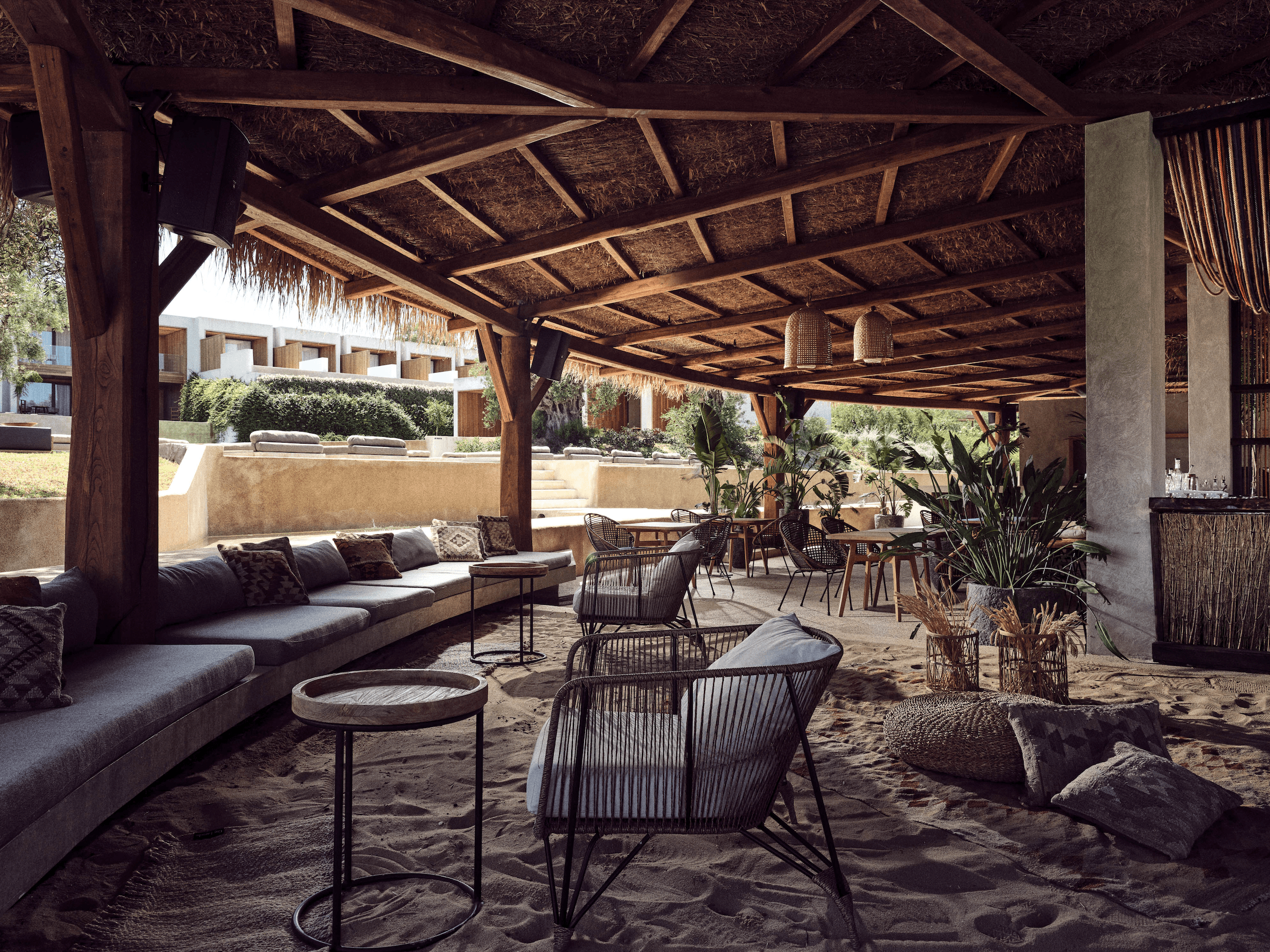 Cocoon Restobar, Europe — Thatched pergola lounge with woven pendant lights, dark upholstered seating, rattan chairs, and sand flooring overlooking manicured gardens.