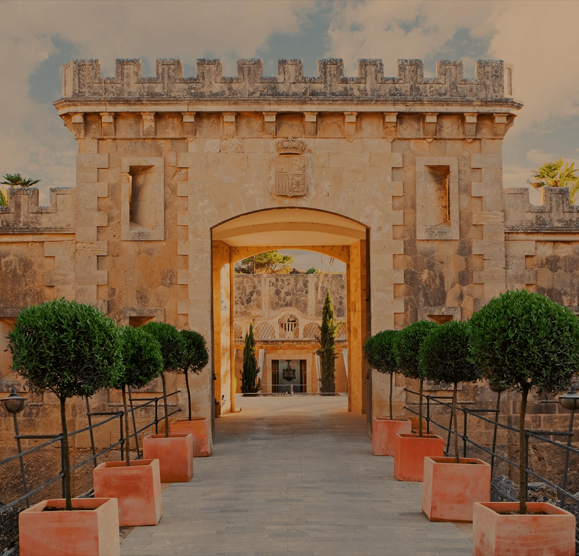 Cap Rocat, Balearic Islands — Fortified stone gateway with barrel-vaulted archway, topiary trees in terracotta planters, Mediterranean courtyard architecture.