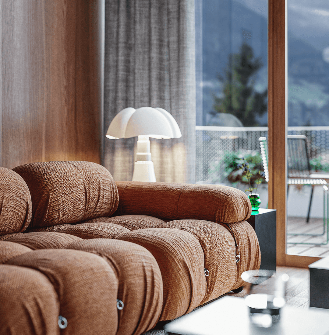 The Cōmodo — Cōmodo, Europe — Sculptural brown upholstered sofa with mushroom lamp beside floor-to-ceiling windows overlooking mountain landscape and terrace.