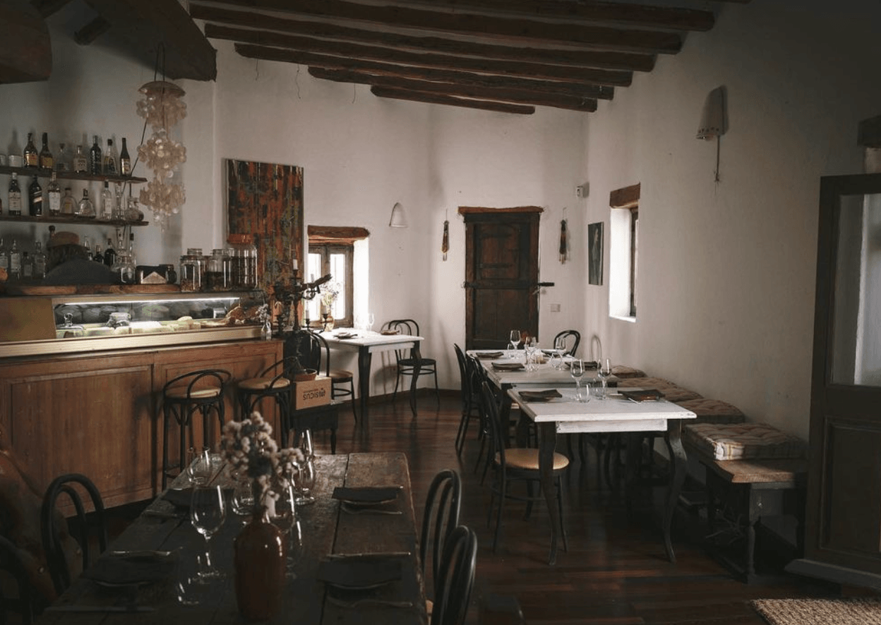 Ibiza Food Studio, Balearic Islands, Spain — Rustic dining room with exposed wooden beams, white walls, dark furniture, bar counter with bottles, and natural light from windows.