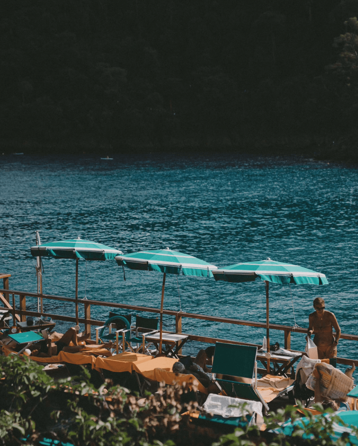 Badni Fiore Beach, Europe — Turquoise umbrellas and canvas loungers line wooden dock overlooking calm teal water, forest backdrop, afternoon light.