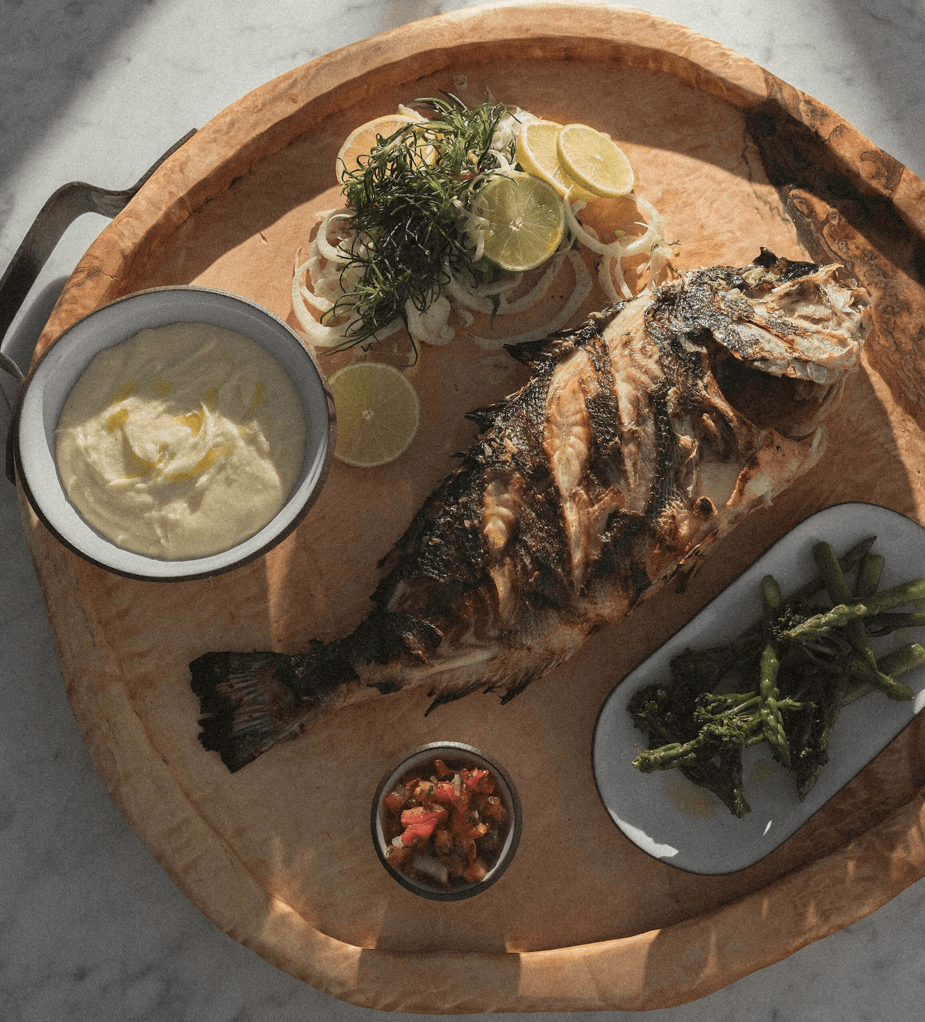Cantine du Faubourg, Europe — Whole grilled fish on wooden board with aioli, charred greens, citrus, and garnishes in natural light.