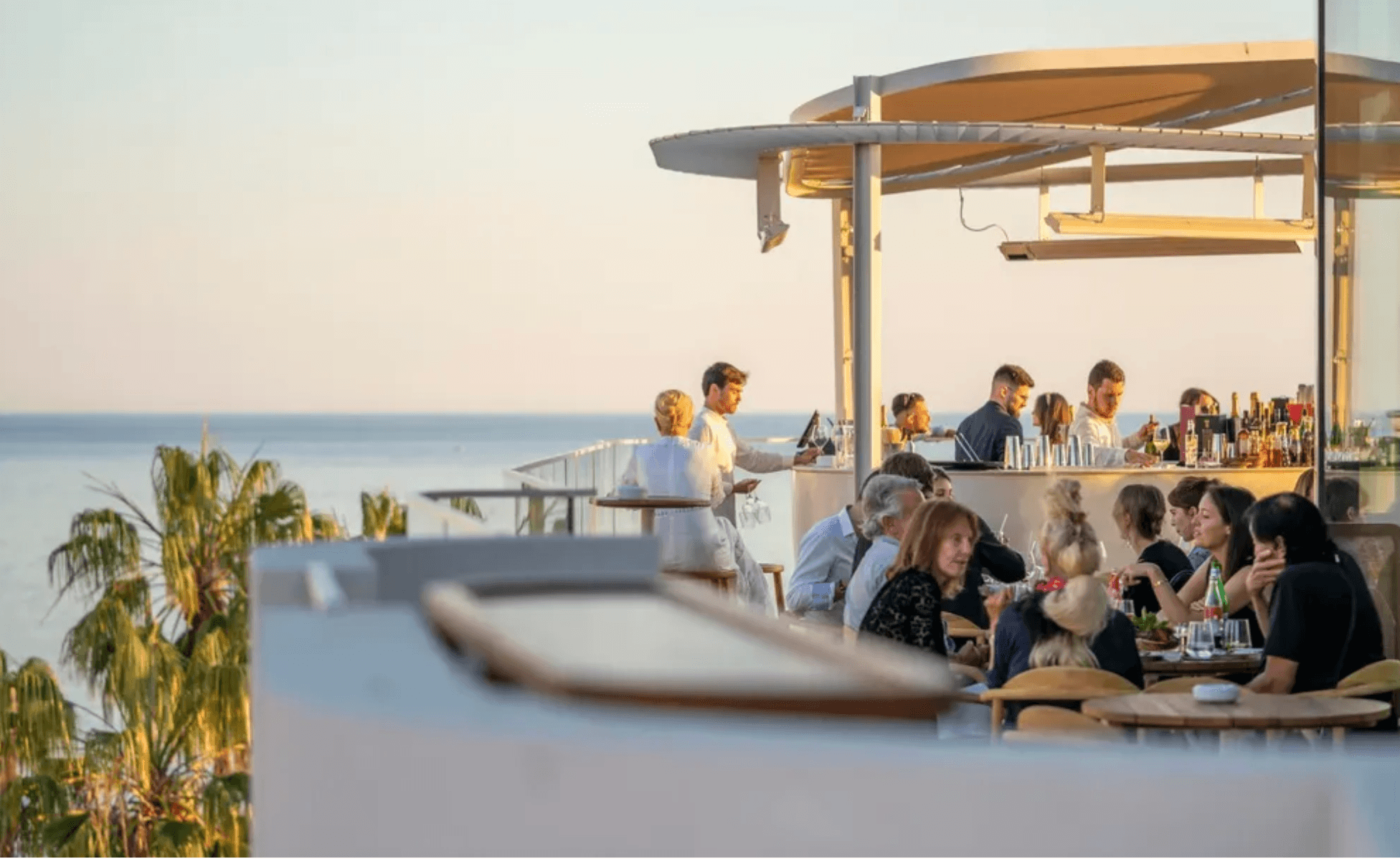 Belle Plage Rooftop Bar, Europe — Sunset-lit rooftop bar with concrete pergola, ocean views, guests dining at tables, palm plants, minimalist hospitality design.