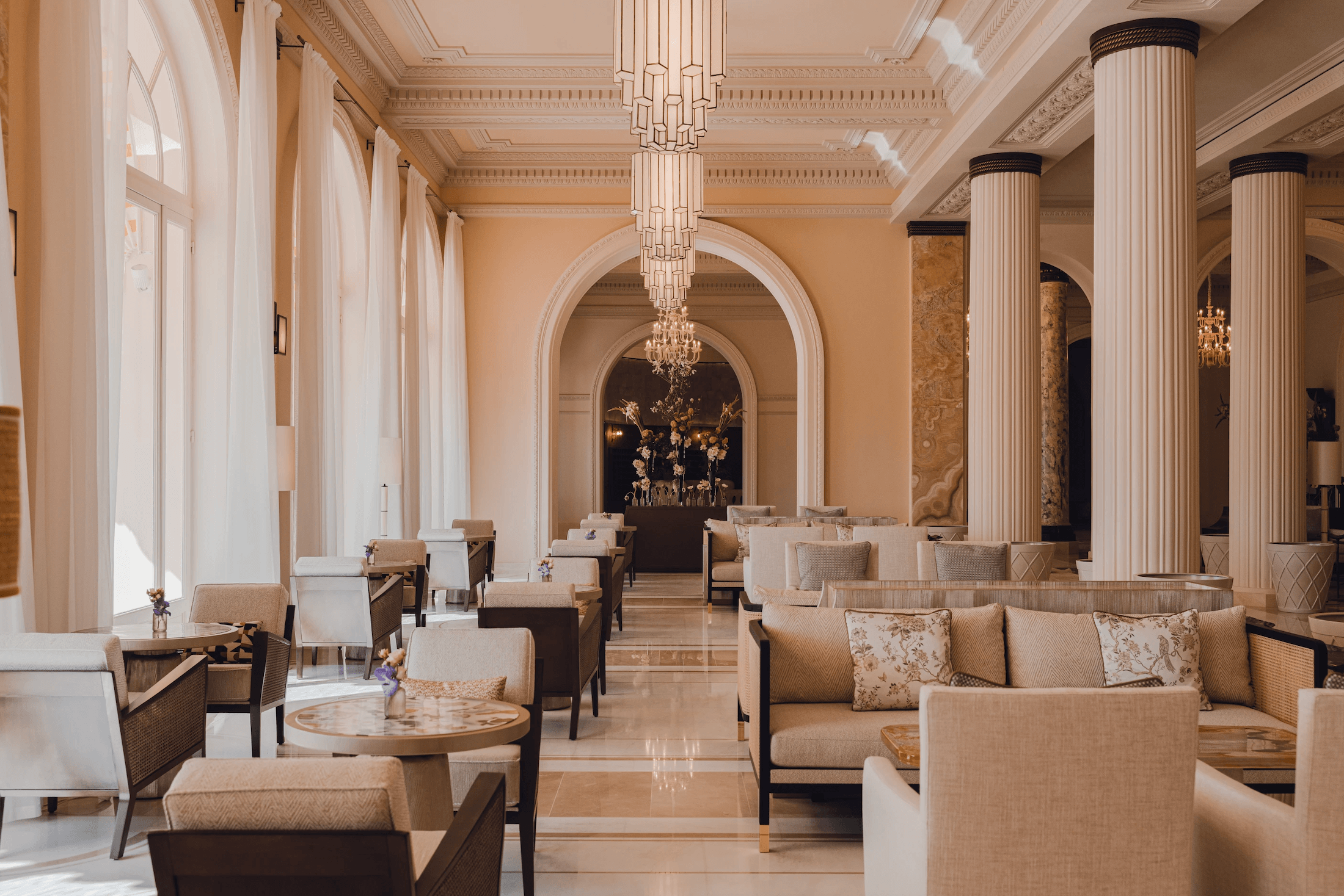 Camélia Tea Lounge, Europe — Neoclassical corridor with fluted columns, cream sectional sofa, geometric inlaid marble floors, crystal chandeliers, and arched doorways bathed in warm natural light.