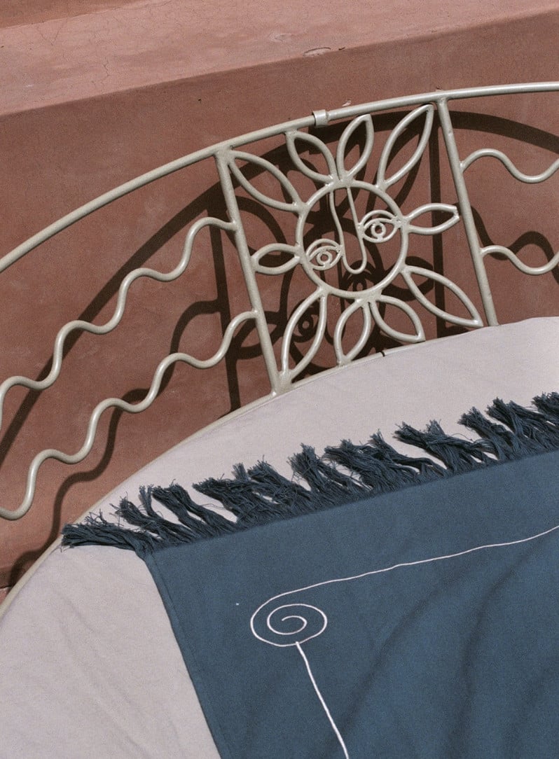 Rosemary, Africa — Ornamental metal headboard with geometric floral motifs and black-fringed linen bedding against terracotta wall.
