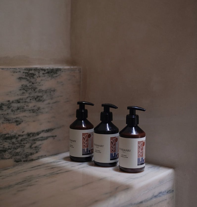 Three dark glass pump bottles with rosemary labels arranged on marble beside textured plaster walls.