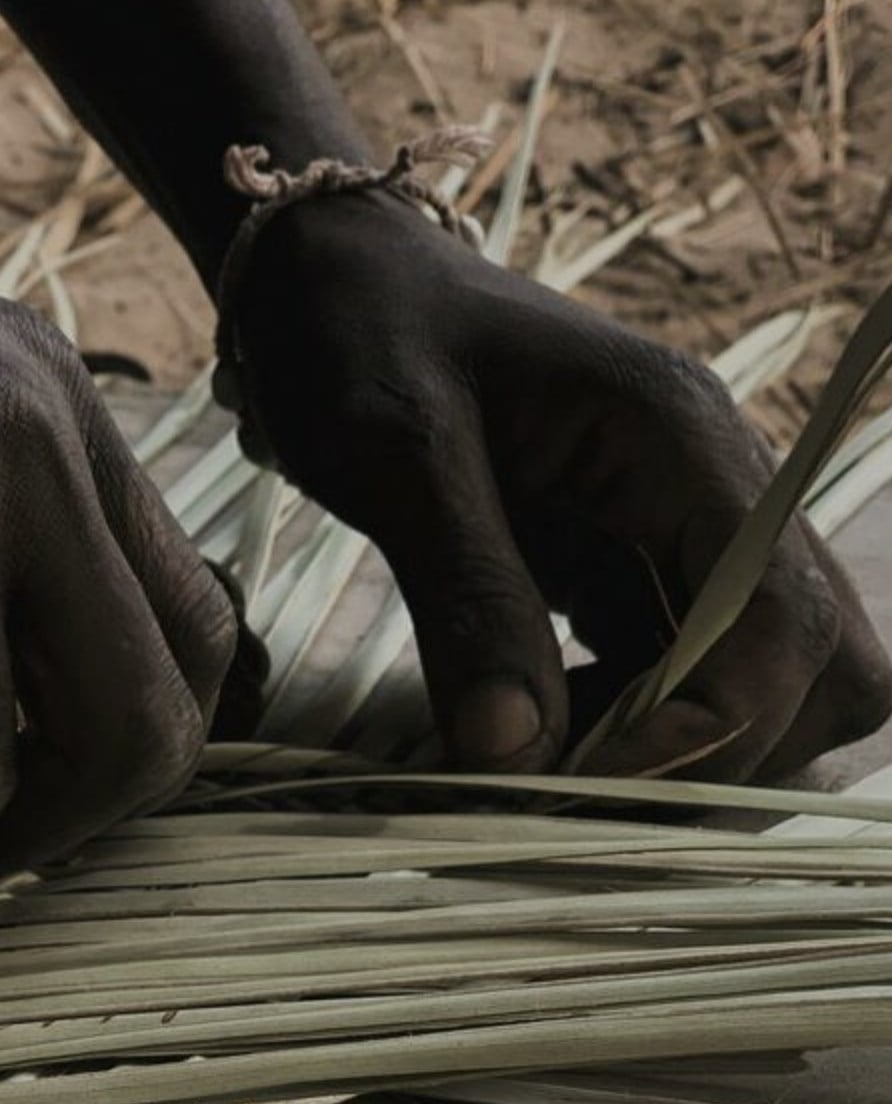 Weaving workshops, Africa — hands working natural fibers on wooden loom with dried palm leaves, artisanal textile craft detail.