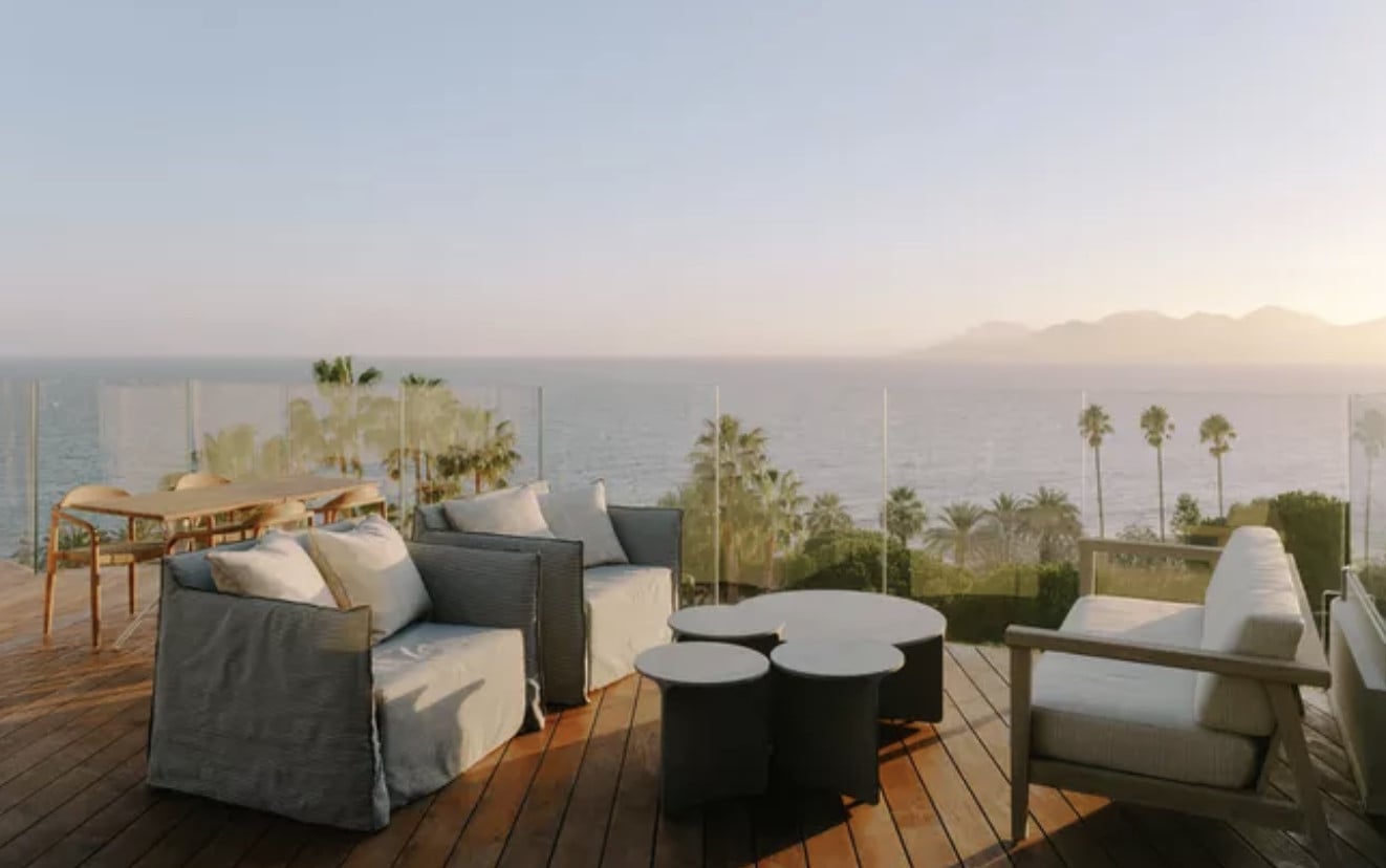 Rooftop Bar, Europe — Sunset terrace with gray upholstered seating, palm trees, sea views, and modern cylindrical side tables on wooden decking.