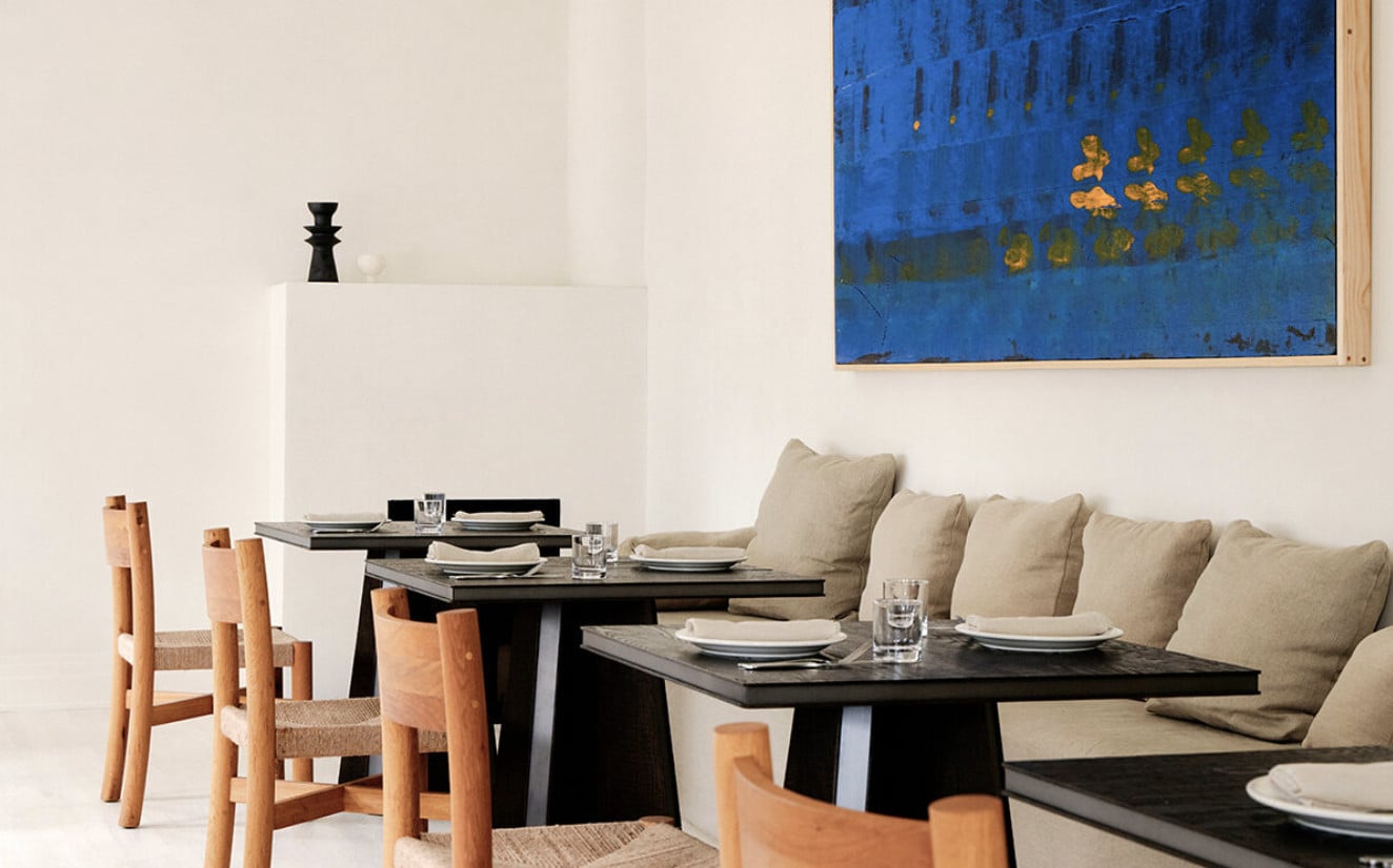 The Garden, North America — Modern dining room with black tables, wooden chairs, beige banquettes, and blue abstract artwork overlooking minimalist interior design.