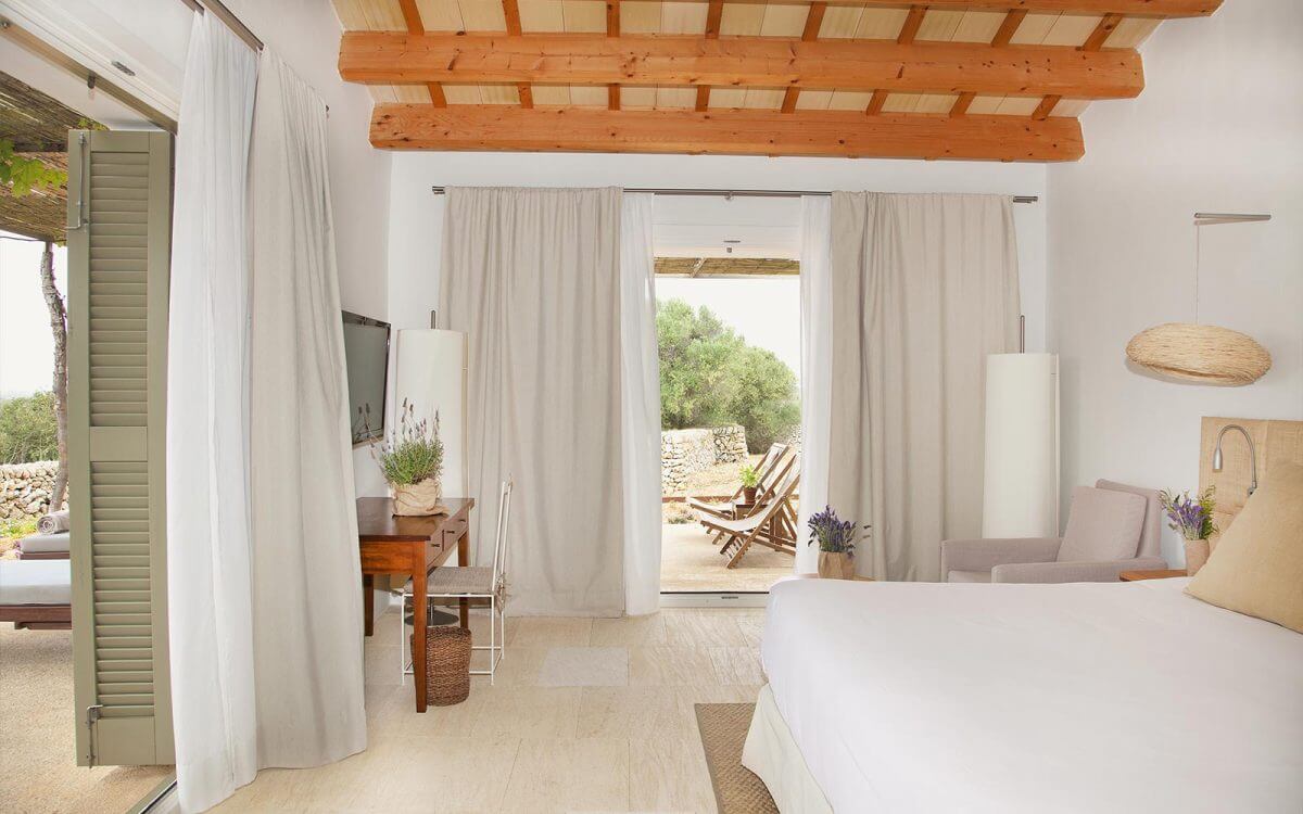 Torralbenc, Balearic Islands — Minimalist bedroom with exposed wooden beams, flowing linen curtains, limestone flooring, and terrace views of Mediterranean landscape.
