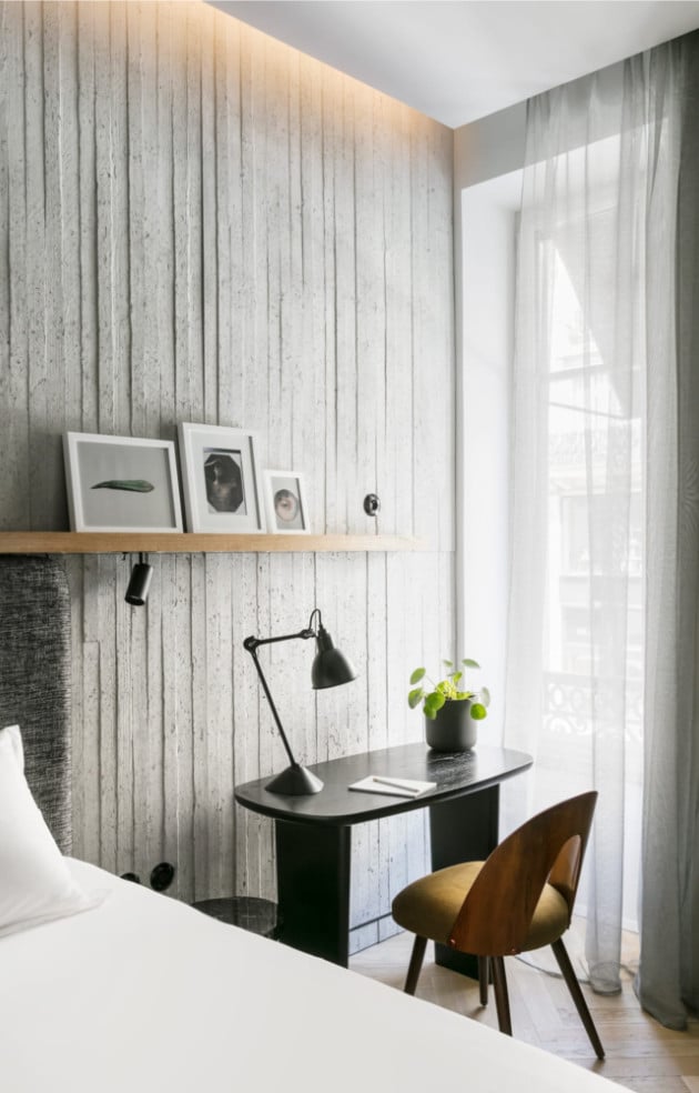 Hôtel National Des Arts, Europe — Modern hotel bedroom nook with whitewashed shiplap walls, floating wooden shelf, black desk, brass chair, and sheer curtains filtering natural light.