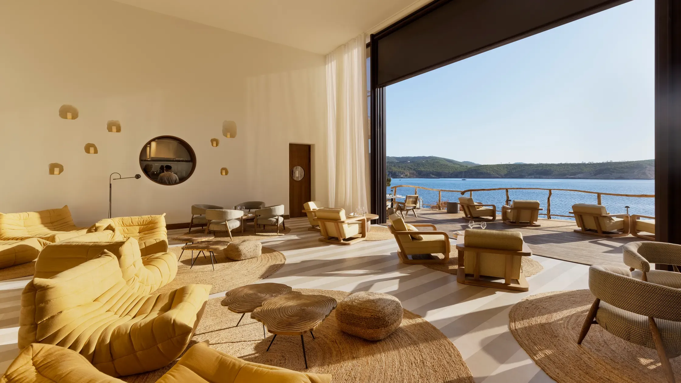 Six Senses Ibiza, Balearic Islands, Spain — Sunlit seafront lounge with golden upholstered furniture, circular mirror, natural materials, and Mediterranean views through floor-to-ceiling glass.