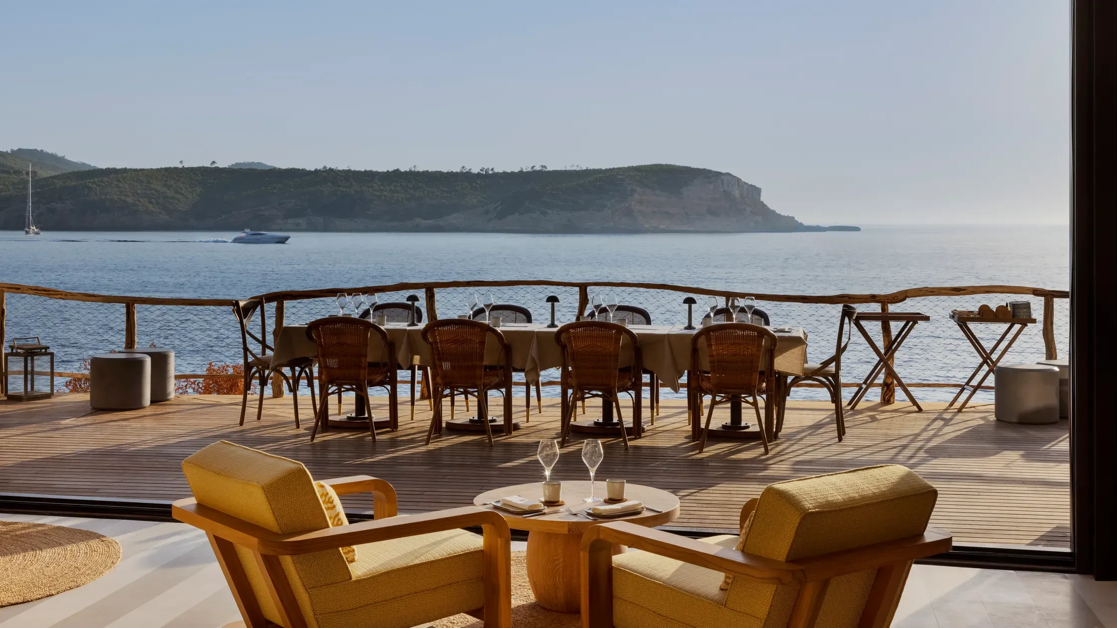 Six Senses Ibiza, Balearic Islands, Spain — Clifftop terrace with rattan dining chairs, wooden deck, and panoramic sea views toward forested headland.