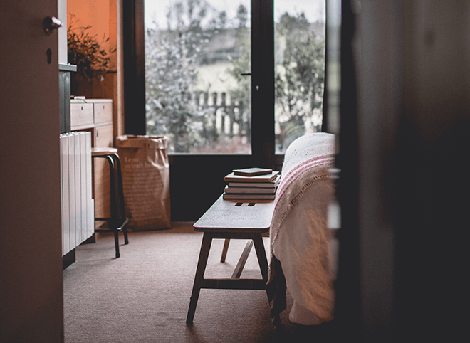 Le Barn, Europe — Minimalist bedroom with wooden chair, stacked books, draped textiles, large windows overlooking countryside architecture and tree-lined landscape in natural light.
