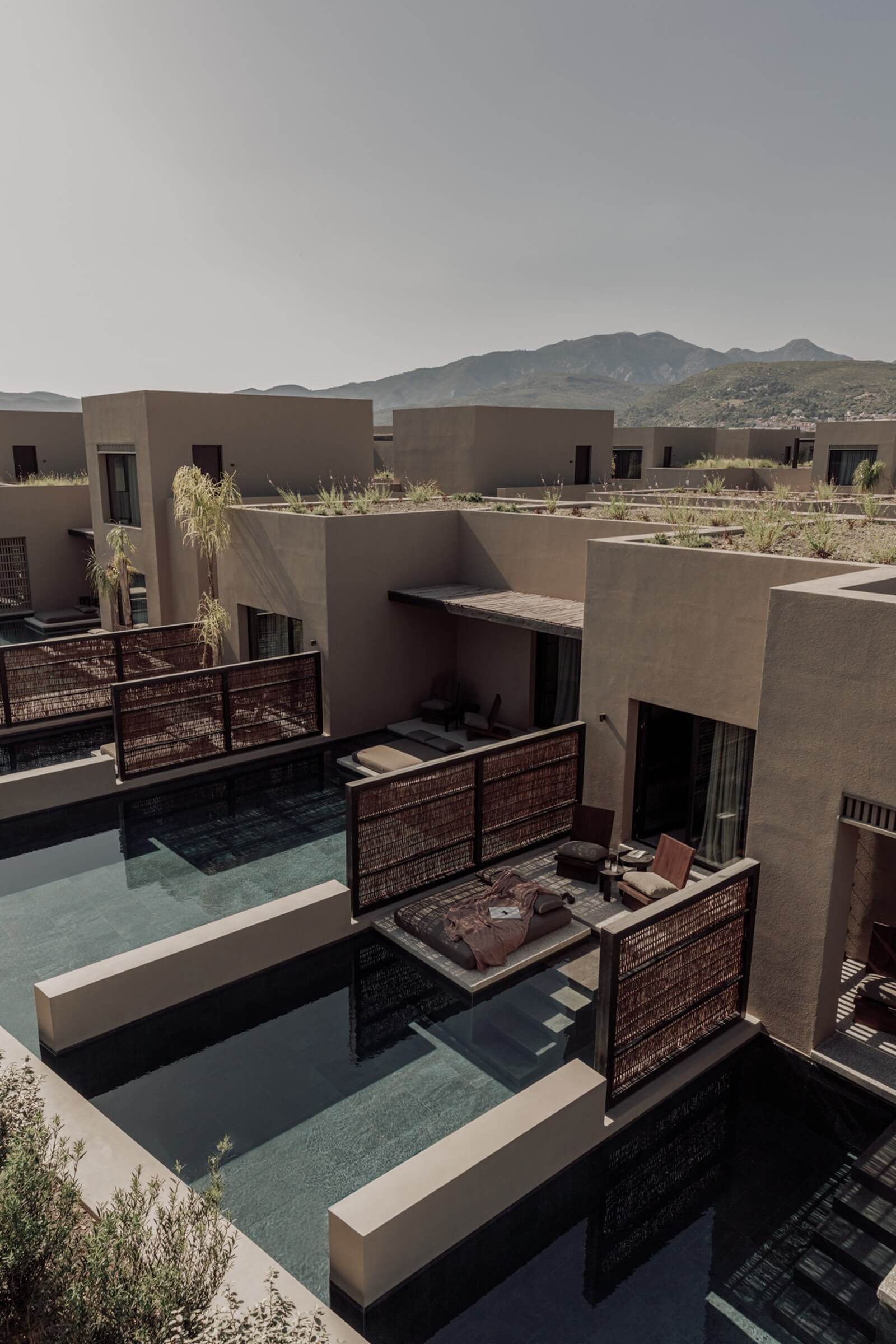 Casa Cook Samos, Greece — Minimalist courtyard with geometric concrete volumes, dark lattice screens, reflecting pools, and mountain views under overcast sky.