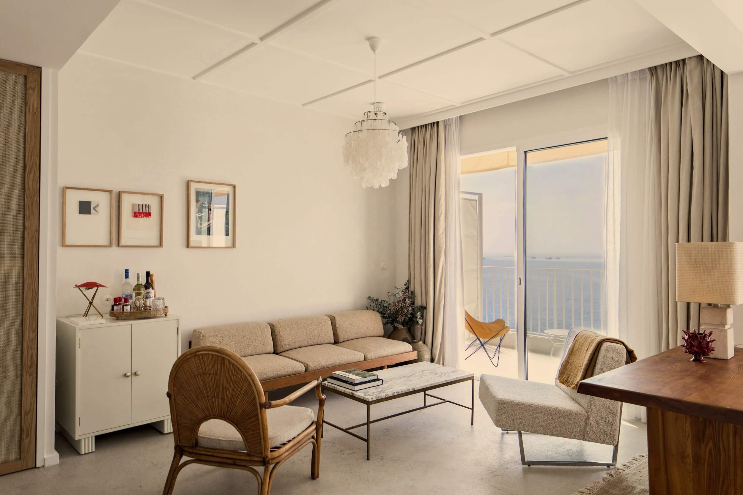 Les Roches Rouges, Europe — Minimalist coastal living room with teak furniture, linen sofa, pendant lighting, and sea views from floor-to-ceiling balcony doors.