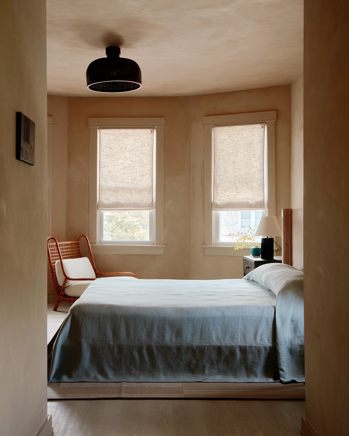 The James Bradley, North America — Minimalist bedroom with linen roller shades, terracotta chair, black pendant light, and blue-grey bedding flooded with natural window light.