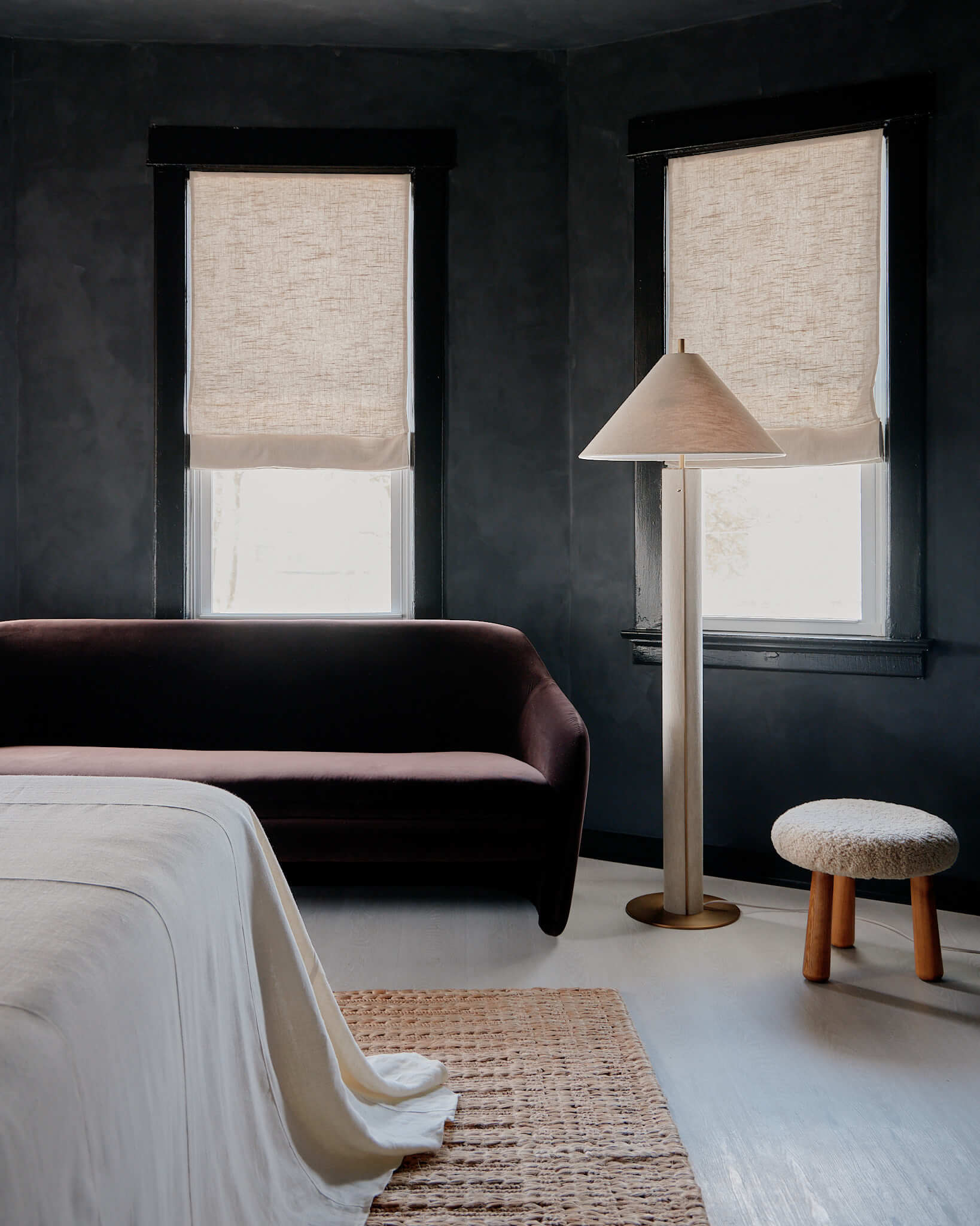 The James Bradley, North America — Contemporary hotel bedroom with charcoal walls, linen roman shades, burgundy curved sofa, natural light, minimalist floor lamp, and neutral textiles.