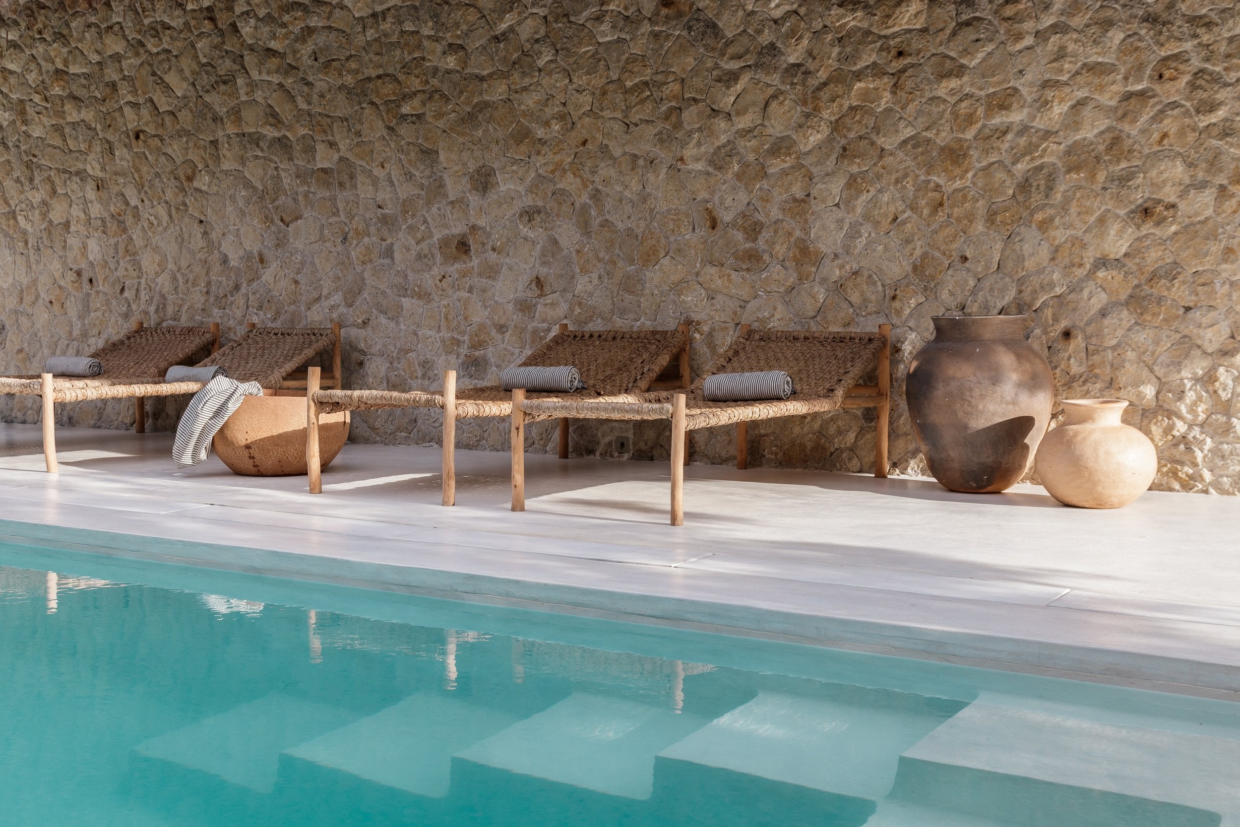 Sussurro, Africa — Poolside terrace with woven lounge chairs, clay vessels, and textured stone wall in warm natural light.