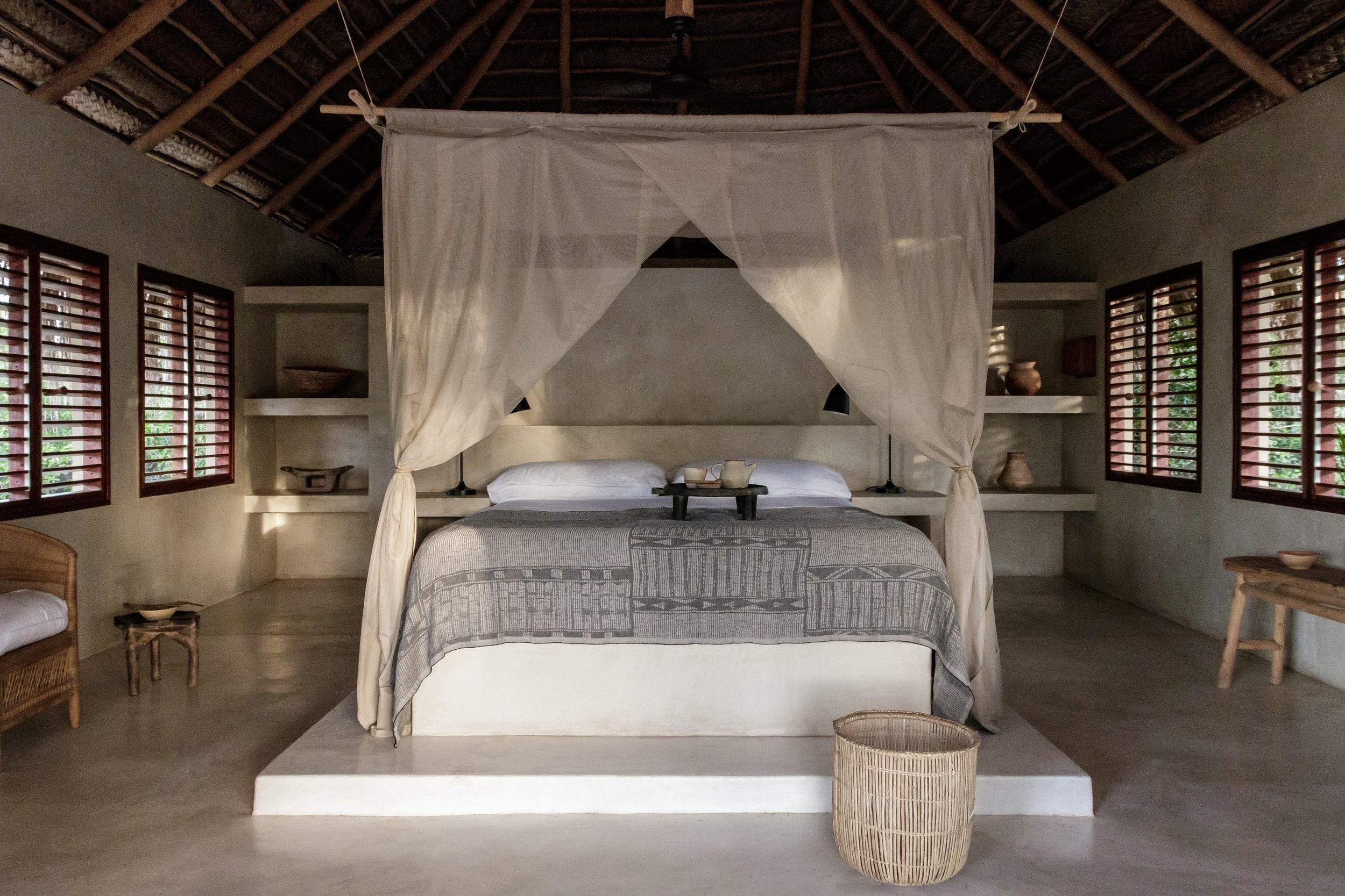 Sussurro, Africa — Minimalist canopy bed with sheer linen curtains in concrete-floor bedroom with louvered windows and exposed timber ceiling.
