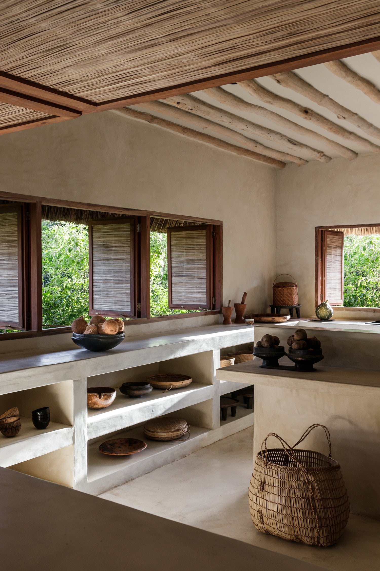 Sussurro, Africa — Minimalist kitchen with concrete counters, open shelving displaying ceramics and woven baskets, exposed wooden beams, and louvered windows opening to lush greenery.