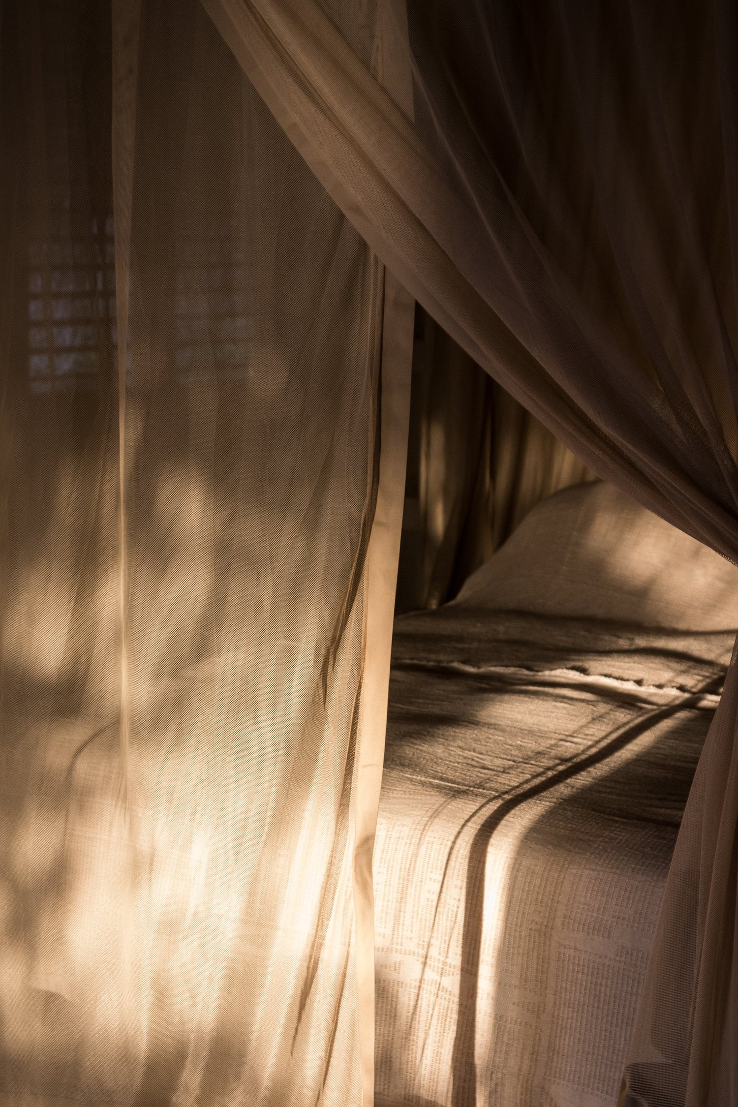 Sussurro, Africa — Sheer linen canopy billows over bed in golden afternoon light, creating intimate draped sanctuary with soft shadows on natural fabric textures.