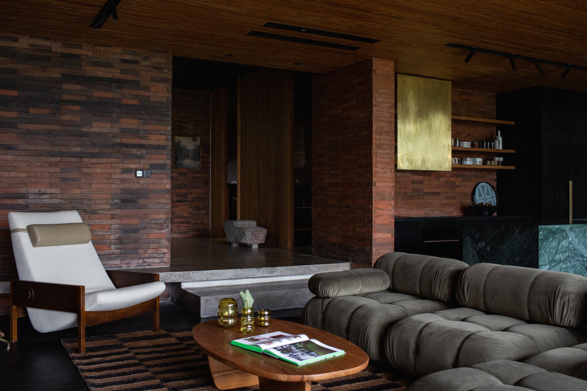 Tegel House, Asia-Pacific — Modernist living room with charcoal sectional sofa, white lounge chair, concrete coffee table, and exposed brick walls beneath wood ceiling.