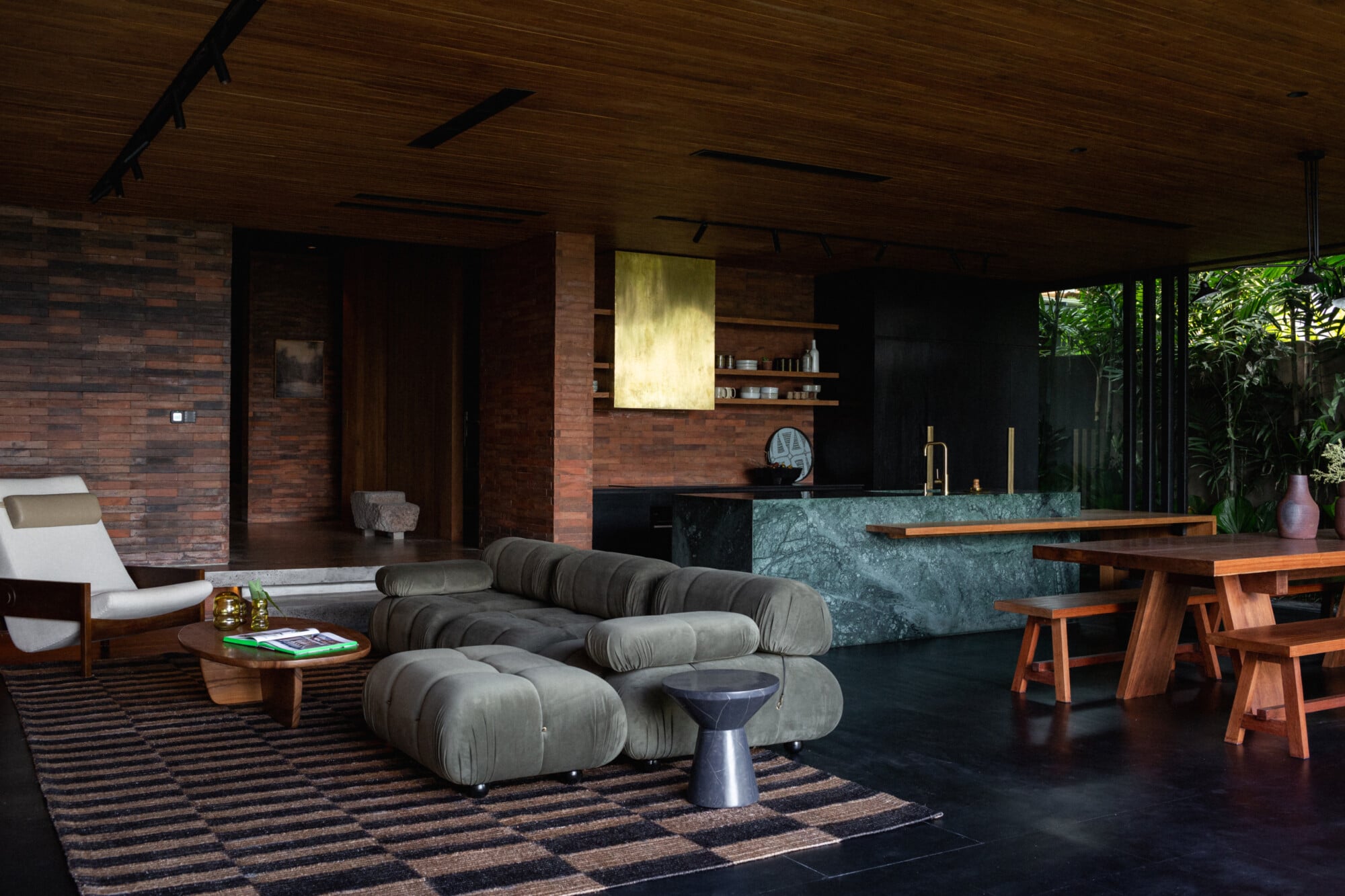 Tegel House, Asia-Pacific — Modern open-plan villa interior with sculptural gray seating, brick walls, dark flooring, wooden dining table, and lush garden views through floor-to-ceiling glass.