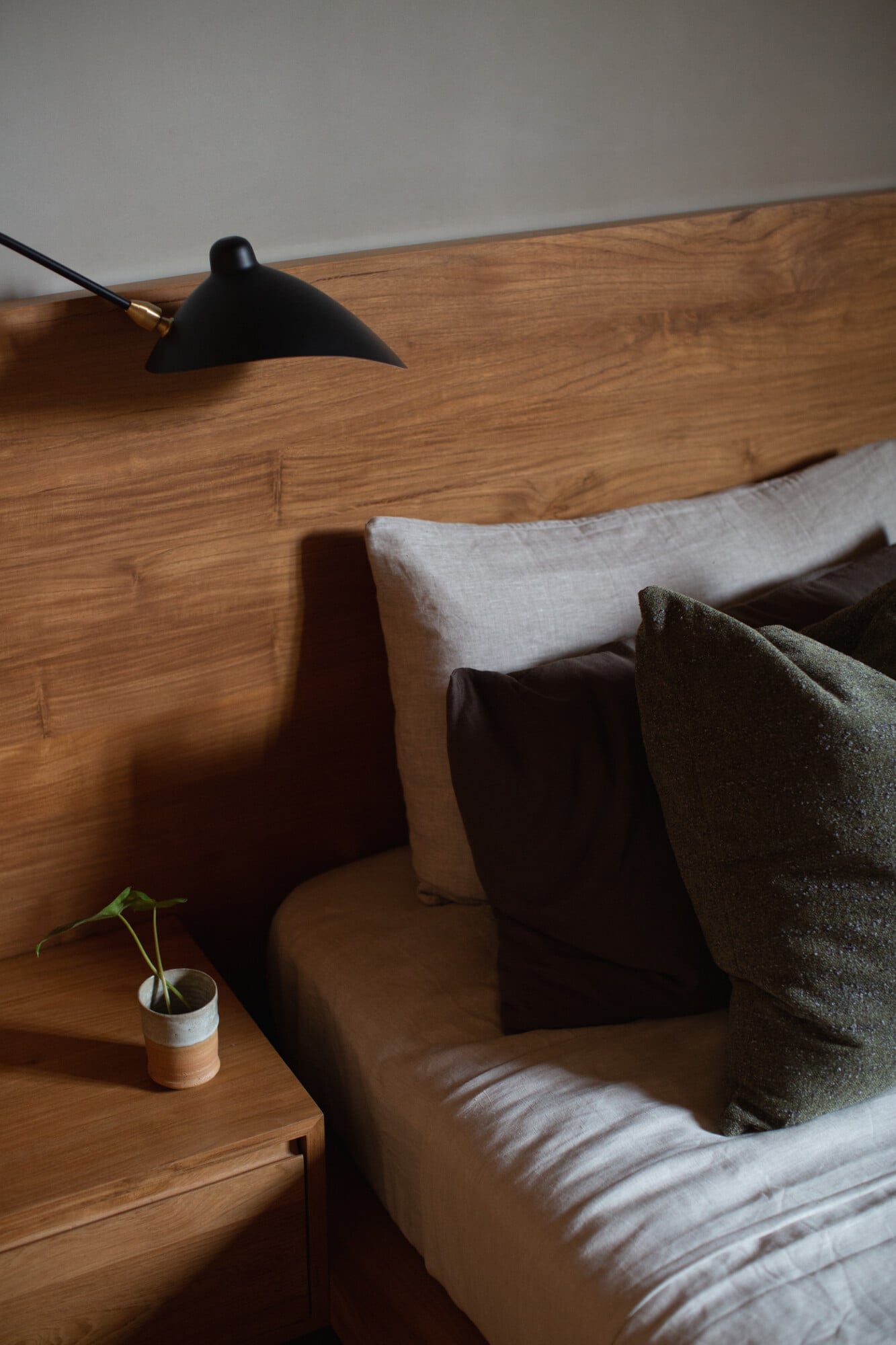 Tegel House, Asia-Pacific — Minimalist bedroom corner with wooden headboard, linen bedding, black task lamp, potted plant, and natural light casting subtle shadows.