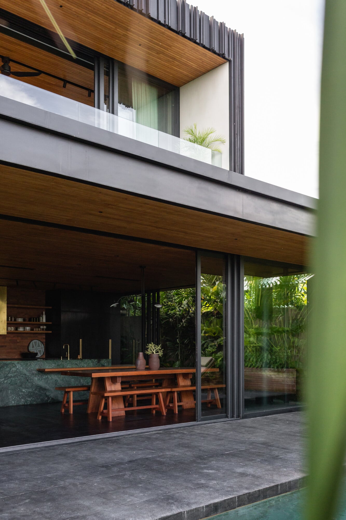 Tegel House, Asia-Pacific — Modern villa interior with timber ceilings, floor-to-ceiling glass doors opening to tropical garden, teak dining furniture, and green-painted courtyard wall.
