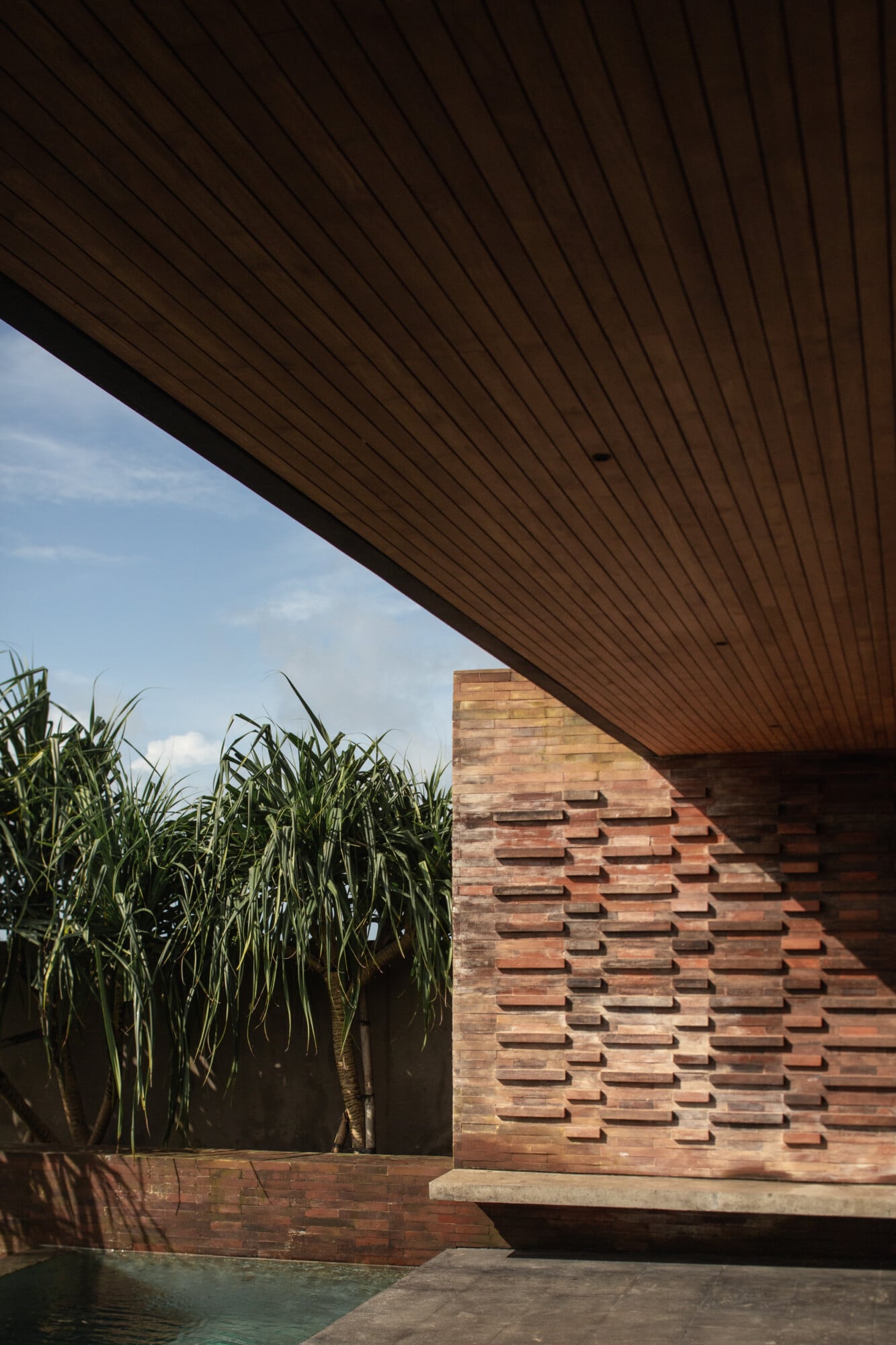 Tegel House, Asia-Pacific — Modernist brick pavilion with staggered facade detailing, wooden soffit overhang, tropical plantings, and reflecting pool beneath clear sky.