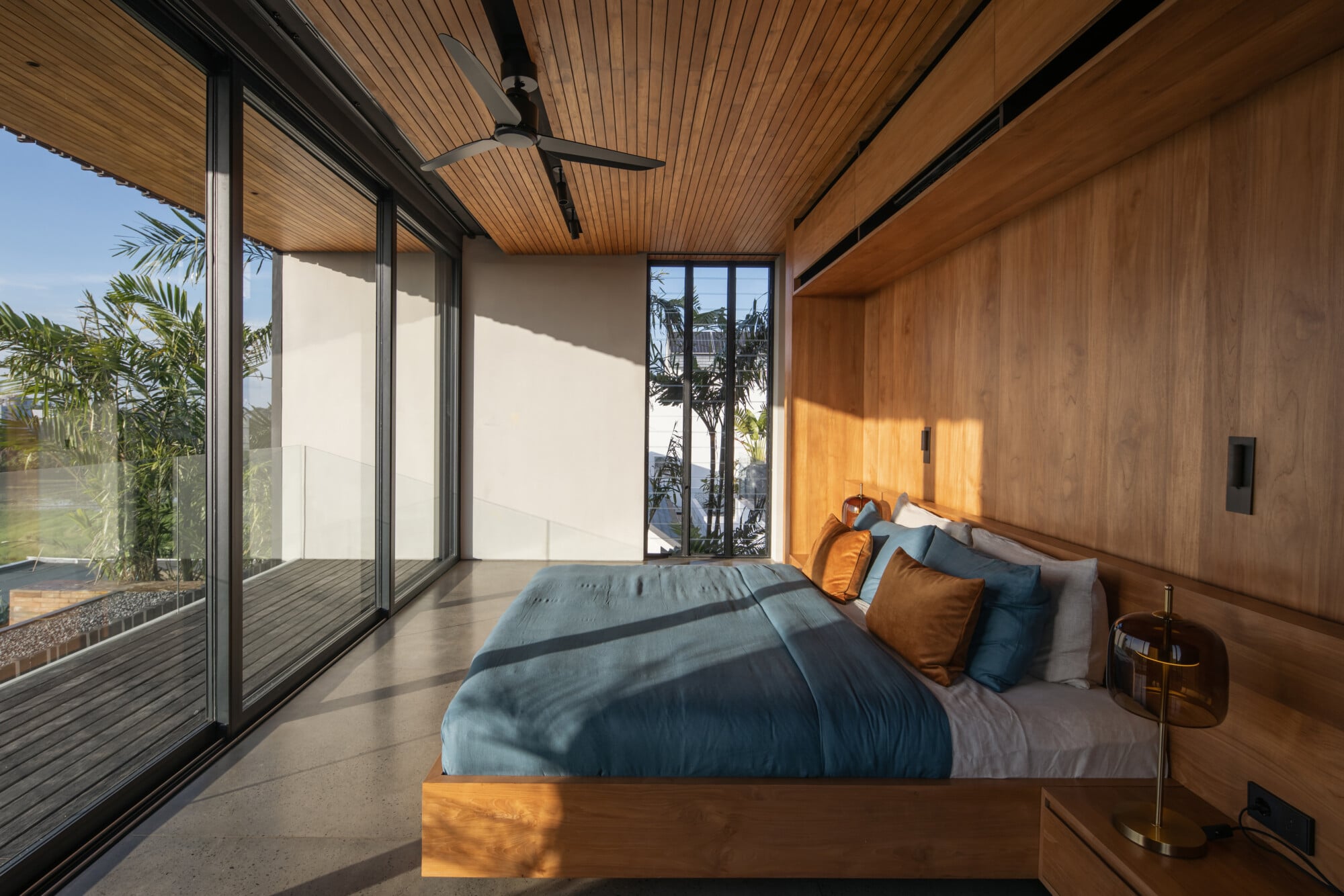 Tegel House, Asia-Pacific — Modern bedroom with warm wood paneling, concrete flooring, blue bedding, floor-to-ceiling glazing overlooking palm trees and water, ceiling fan, morning light.