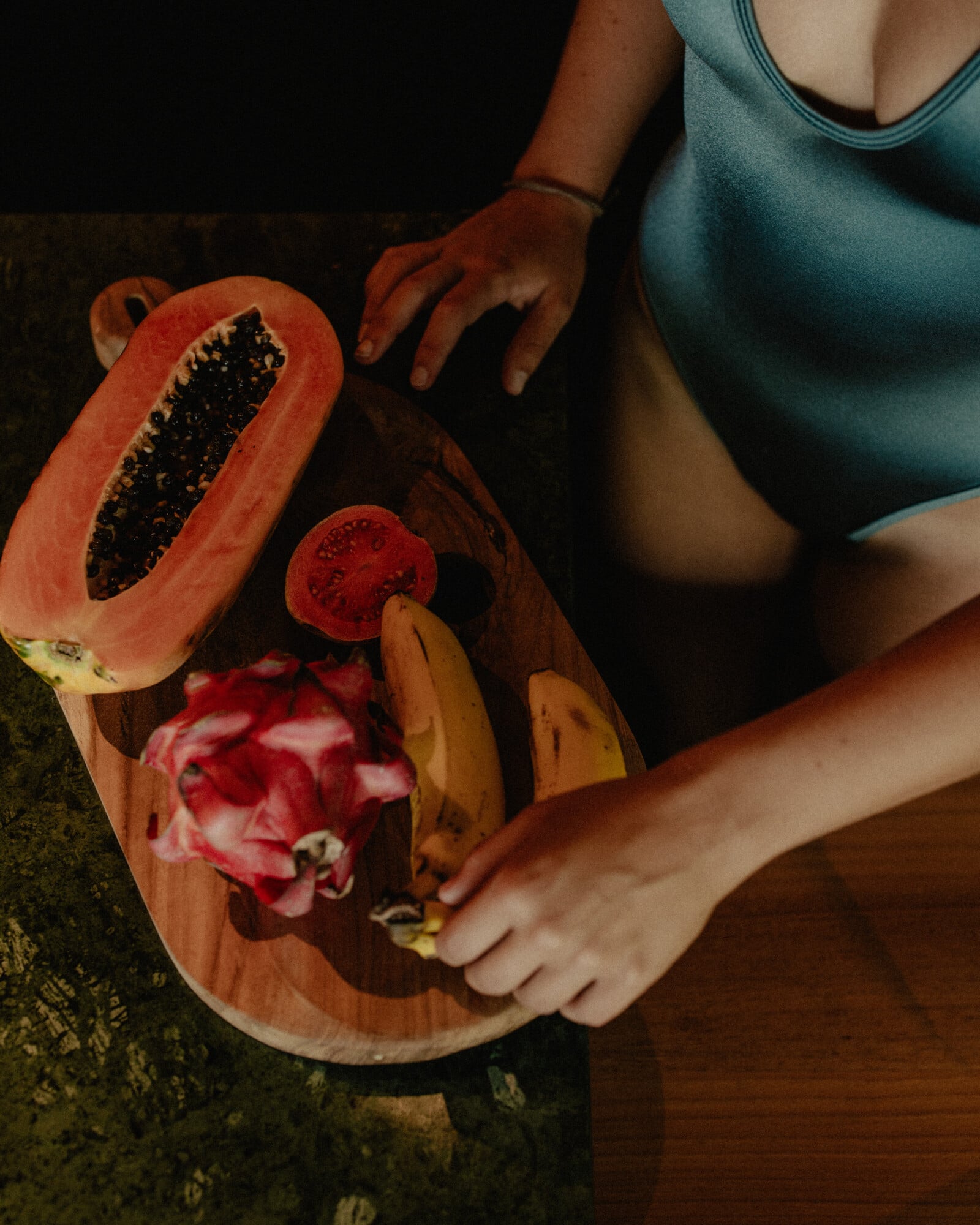 Tegel House, Asia-Pacific — Woman in teal tank top arranging fresh papaya and flowers on wooden board, warm natural light casting shadows on textured surface.