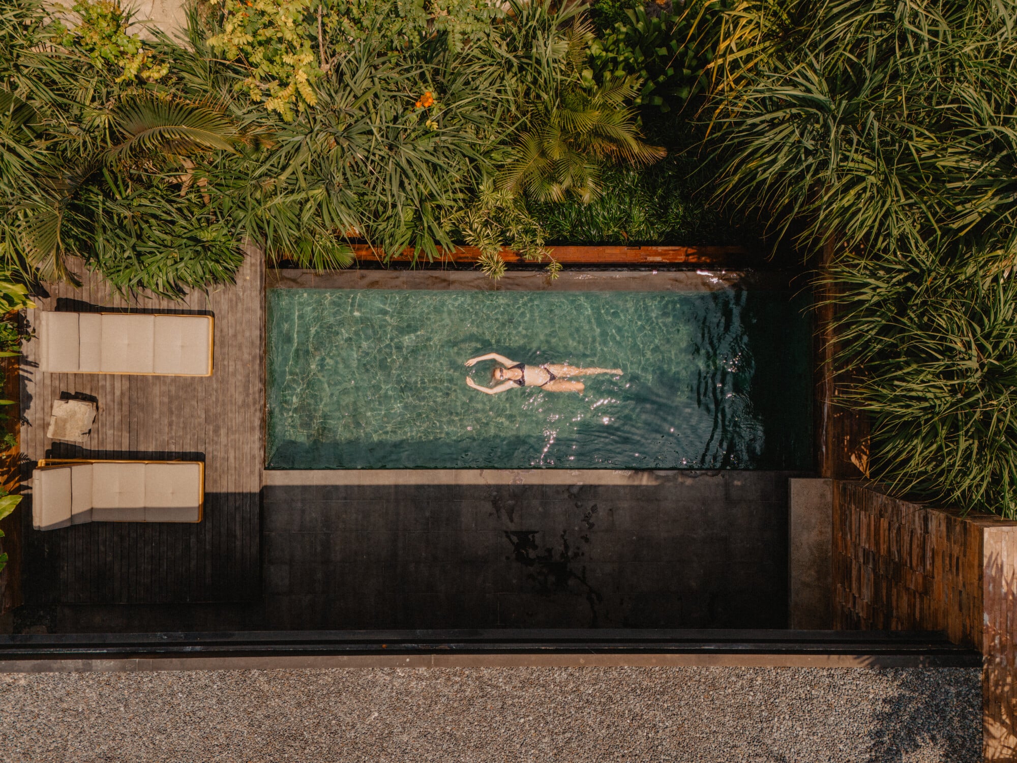 Tegel House, Asia-Pacific — Aerial view of modernist infinity pool with turquoise water, person floating, surrounded by tropical palms and weathered timber decking.